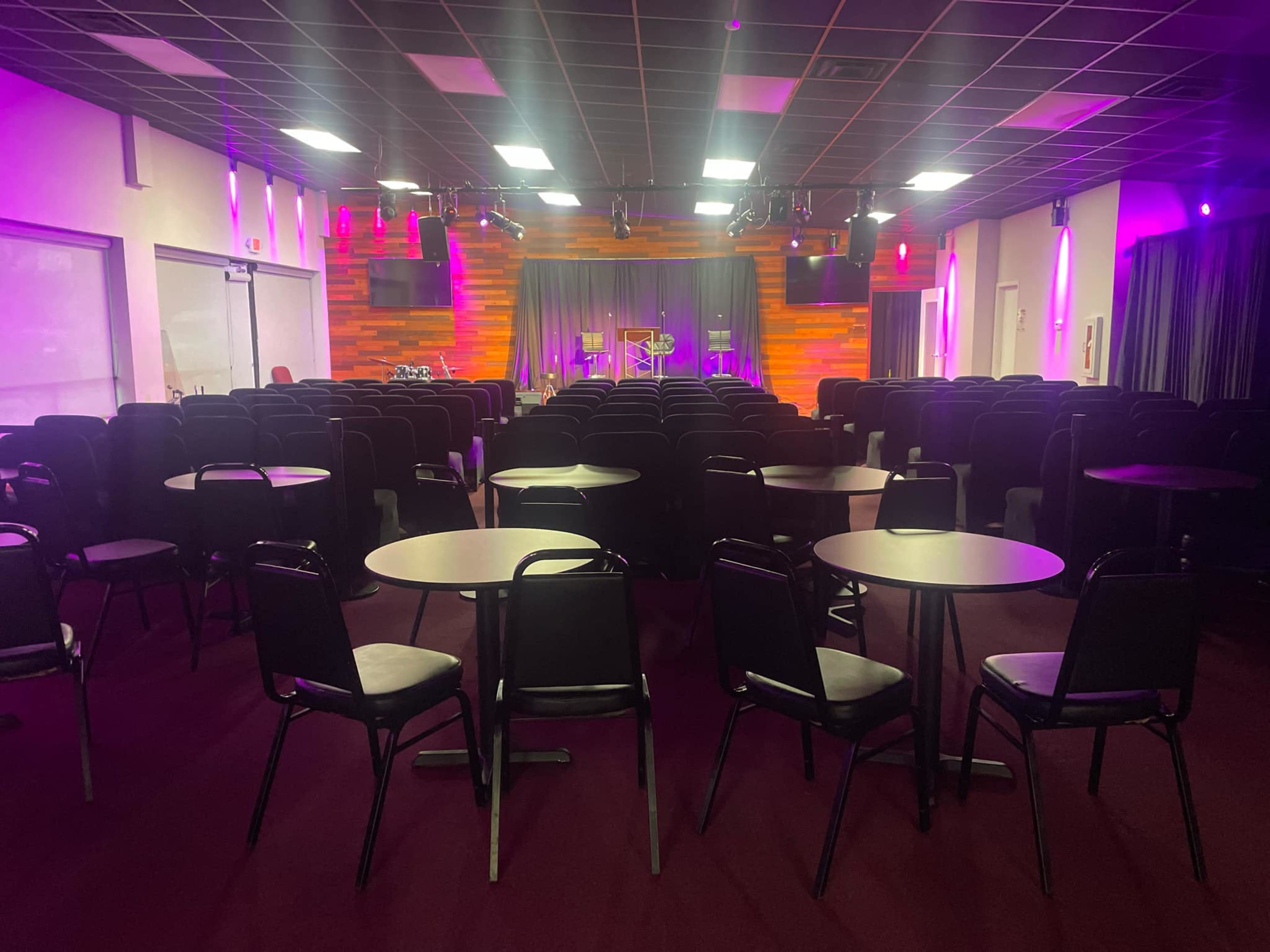 The image shows a seating arrangement in a dimly lit performance venue, featuring rows of round tables and chairs facing a stage with a curtain and lights.