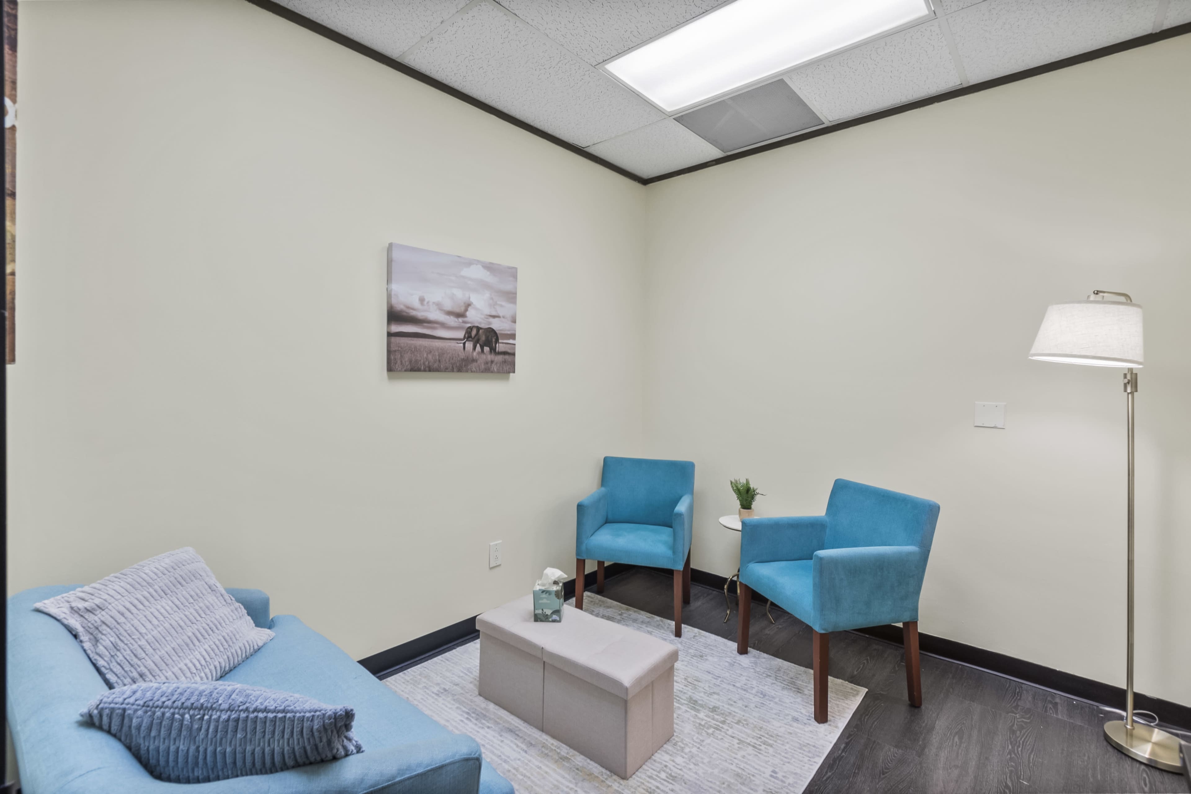 The image shows a small, well-lit room furnished with a blue sofa, two matching chairs, a lamp, and a small coffee table.