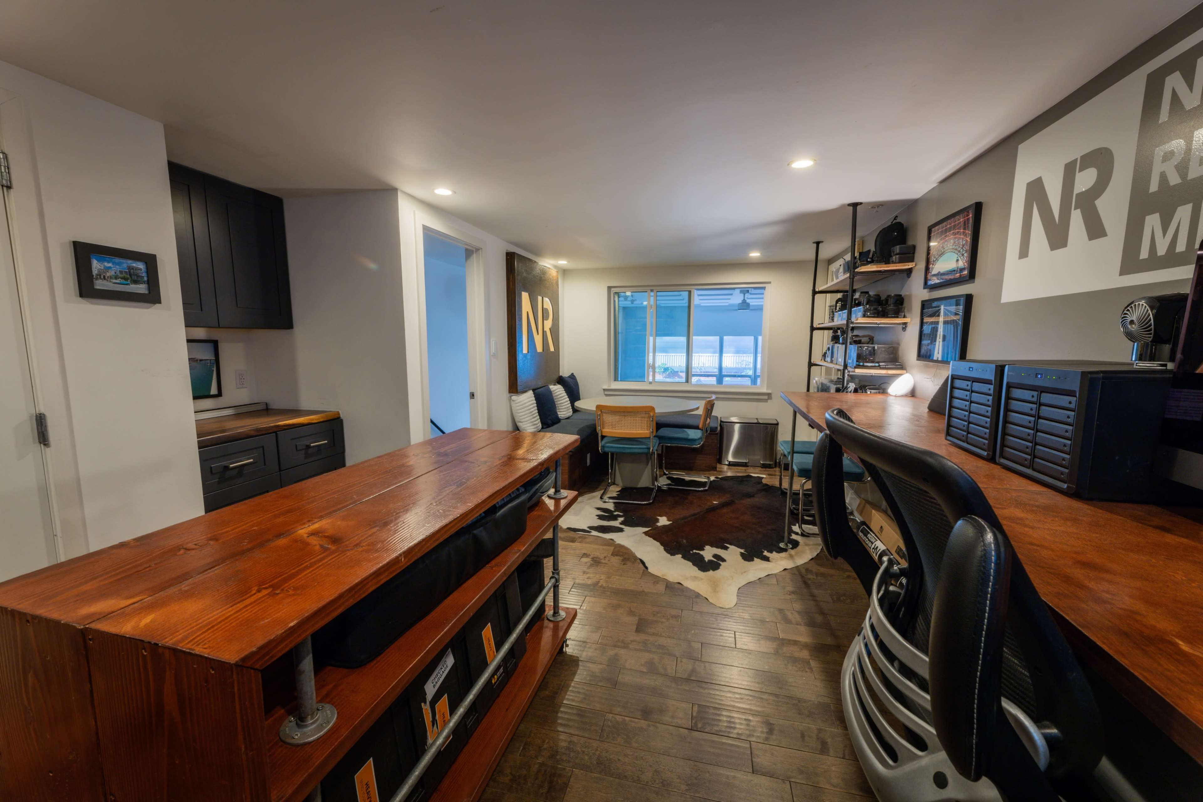 The image shows a modern home office space with a wooden bar counter, a work desk with chairs, and shelves containing various items against a light-colored wall.