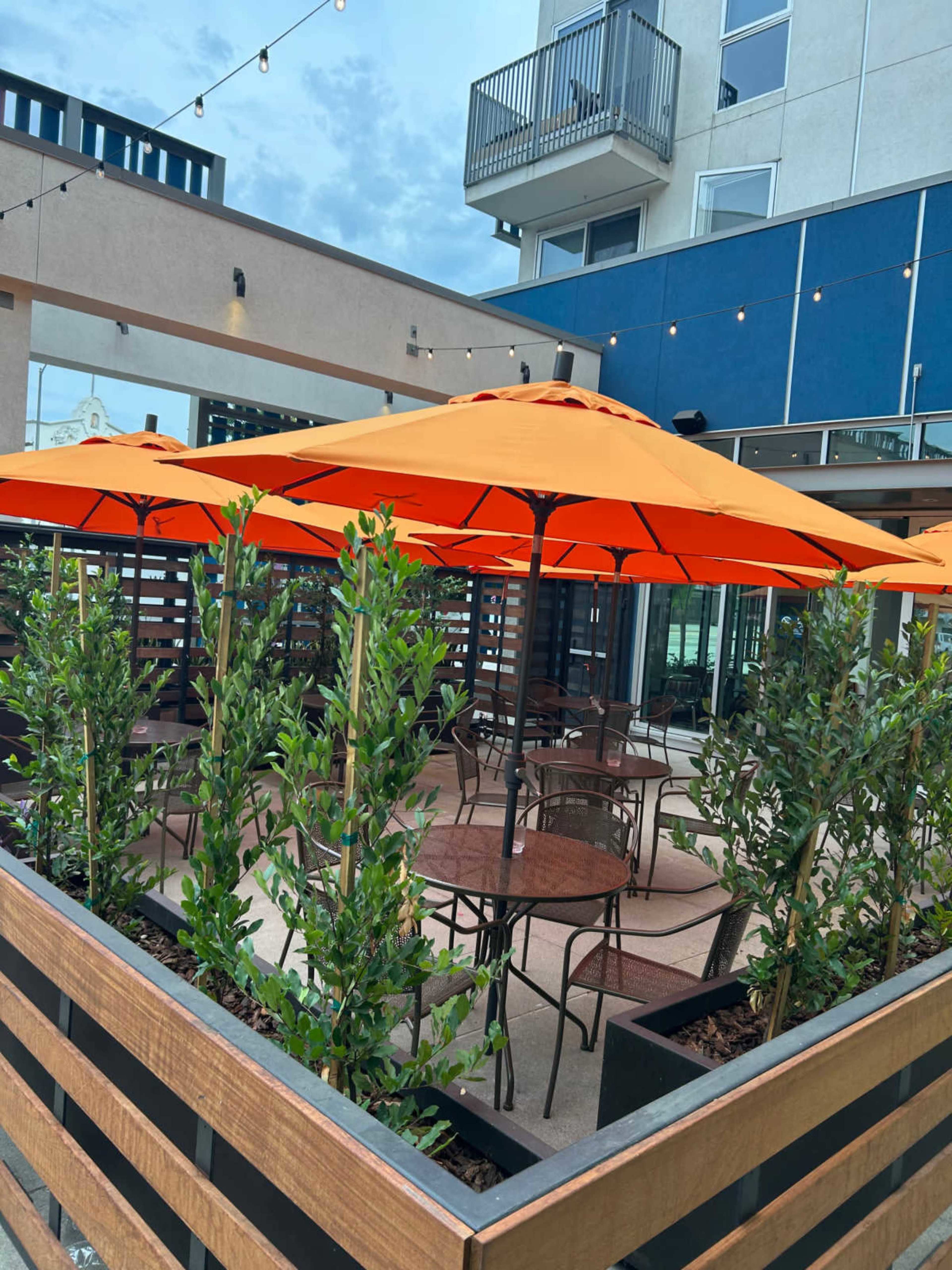 The image depicts an outdoor dining area with orange umbrellas and green plants arranged around metal tables and chairs.