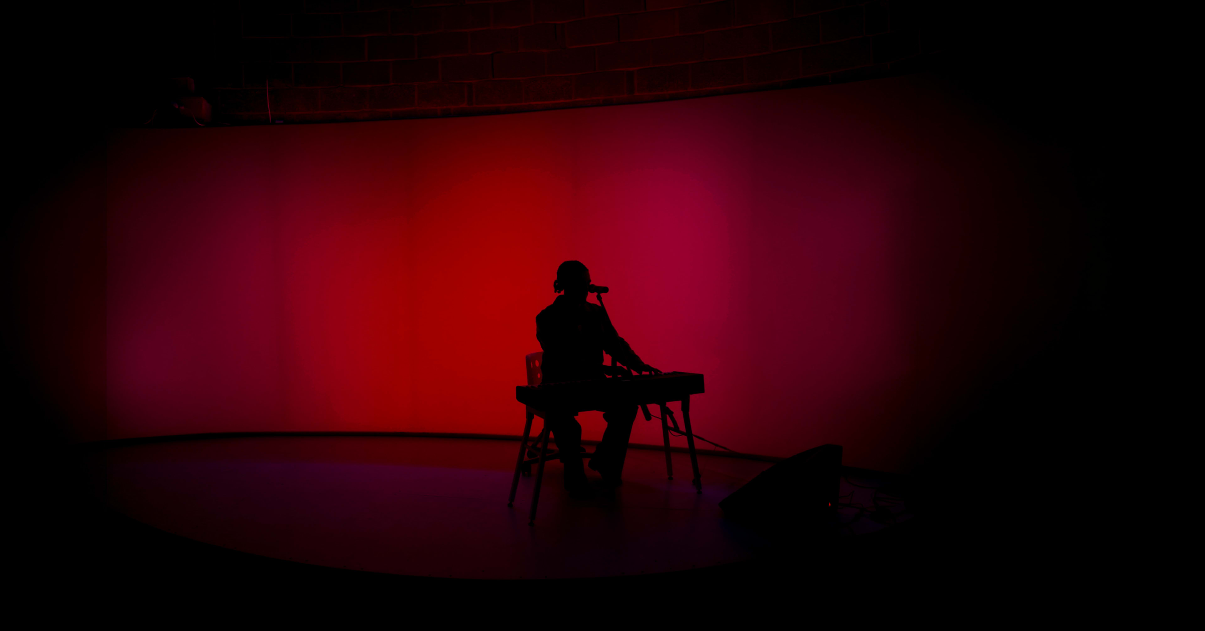 A musician sits at a keyboard in silhouette against a brightly lit pink and red background.