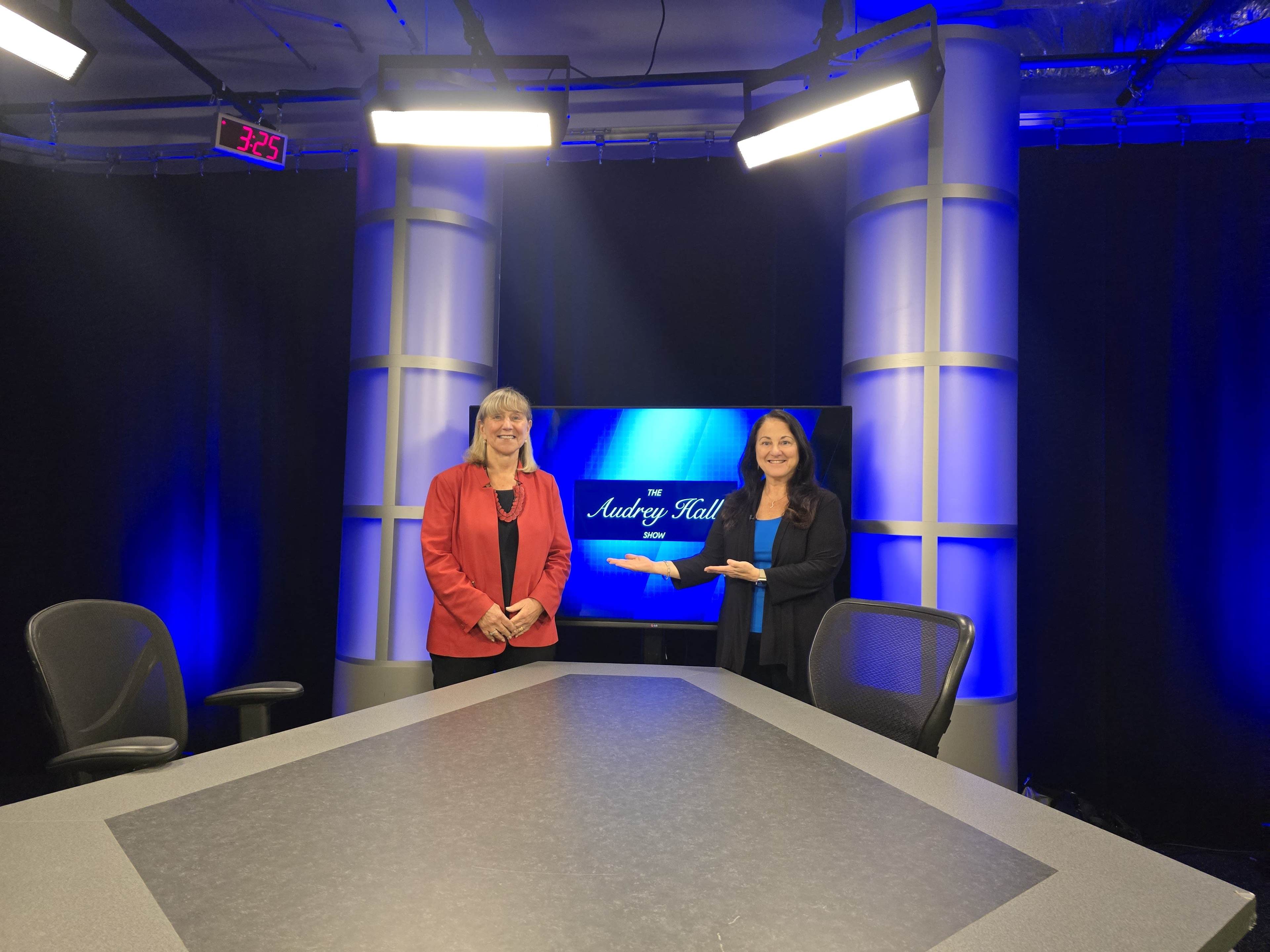 Two women stand in a television studio with a blue-themed backdrop displaying "The Audrey Hall" and a large table in front of them.