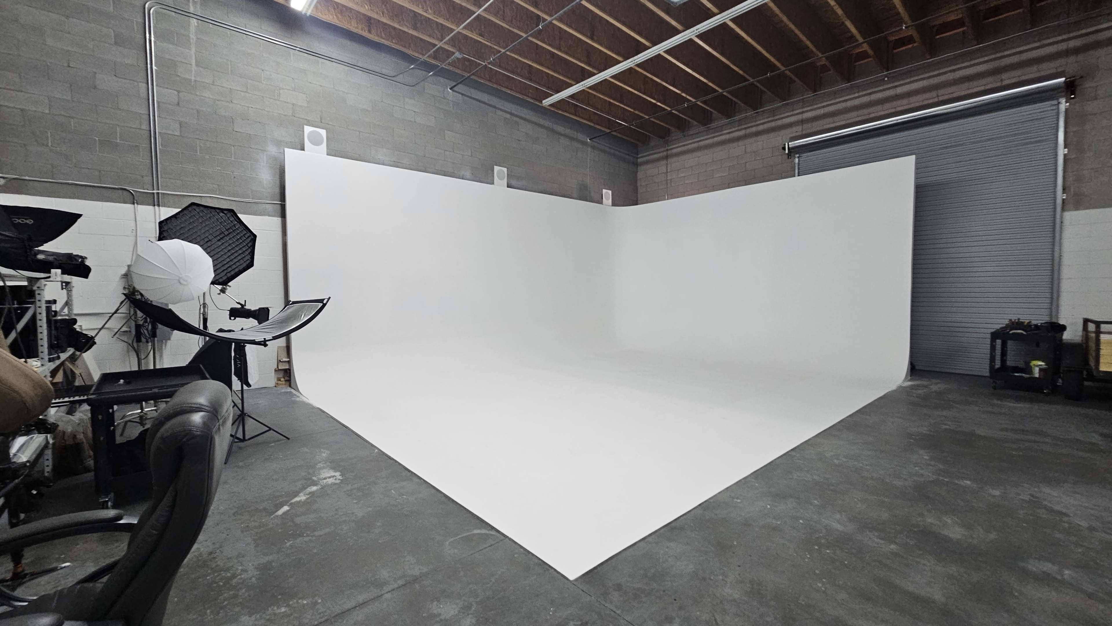 The image shows a photography studio with a large, seamless white backdrop and studio equipment, including lighting gear and a chair.