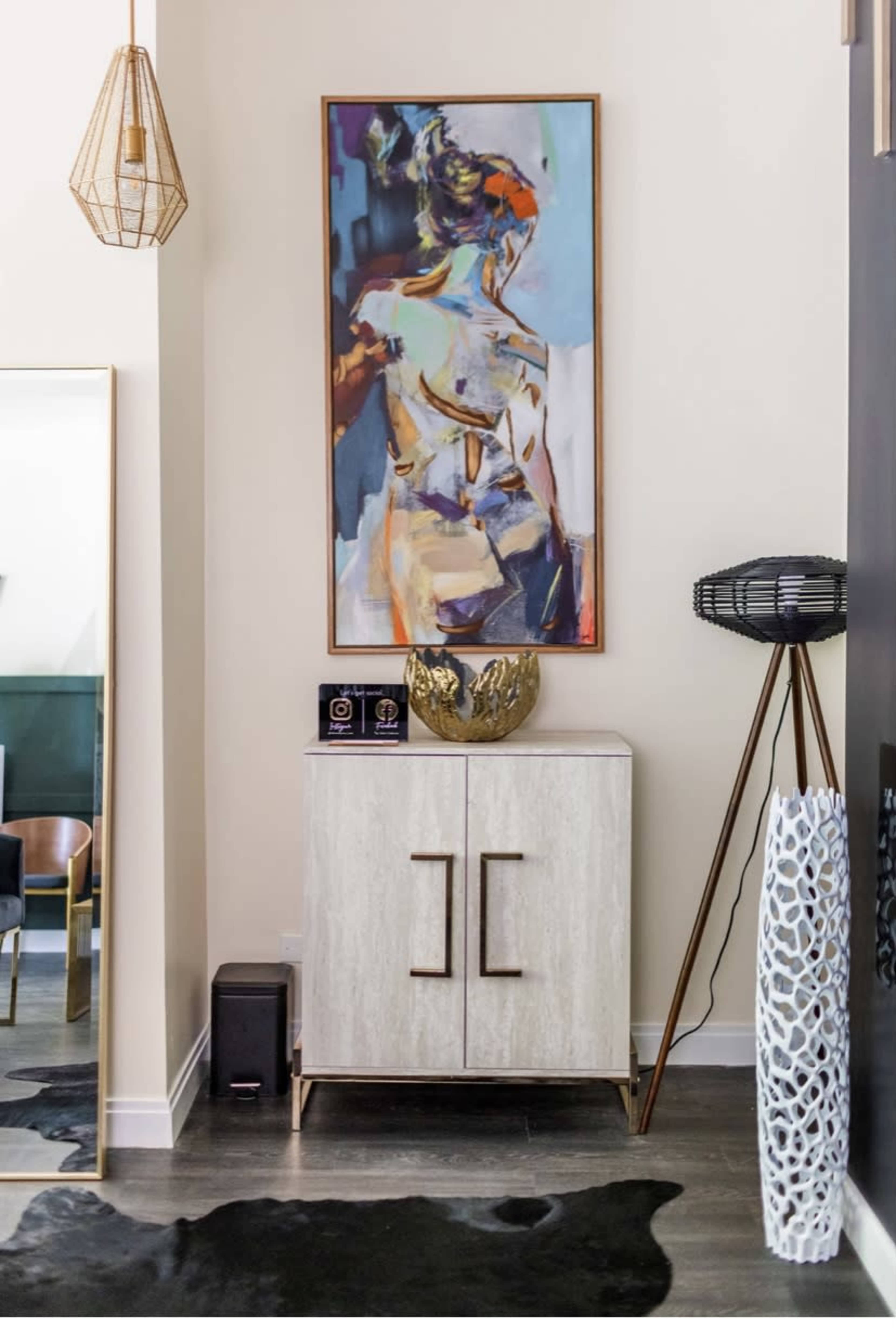 A modern entryway featuring a cabinet with two doors, a colorful framed painting above it, a decorative bowl, and a standing lamp, all accented by a patterned rug and stylish decor.