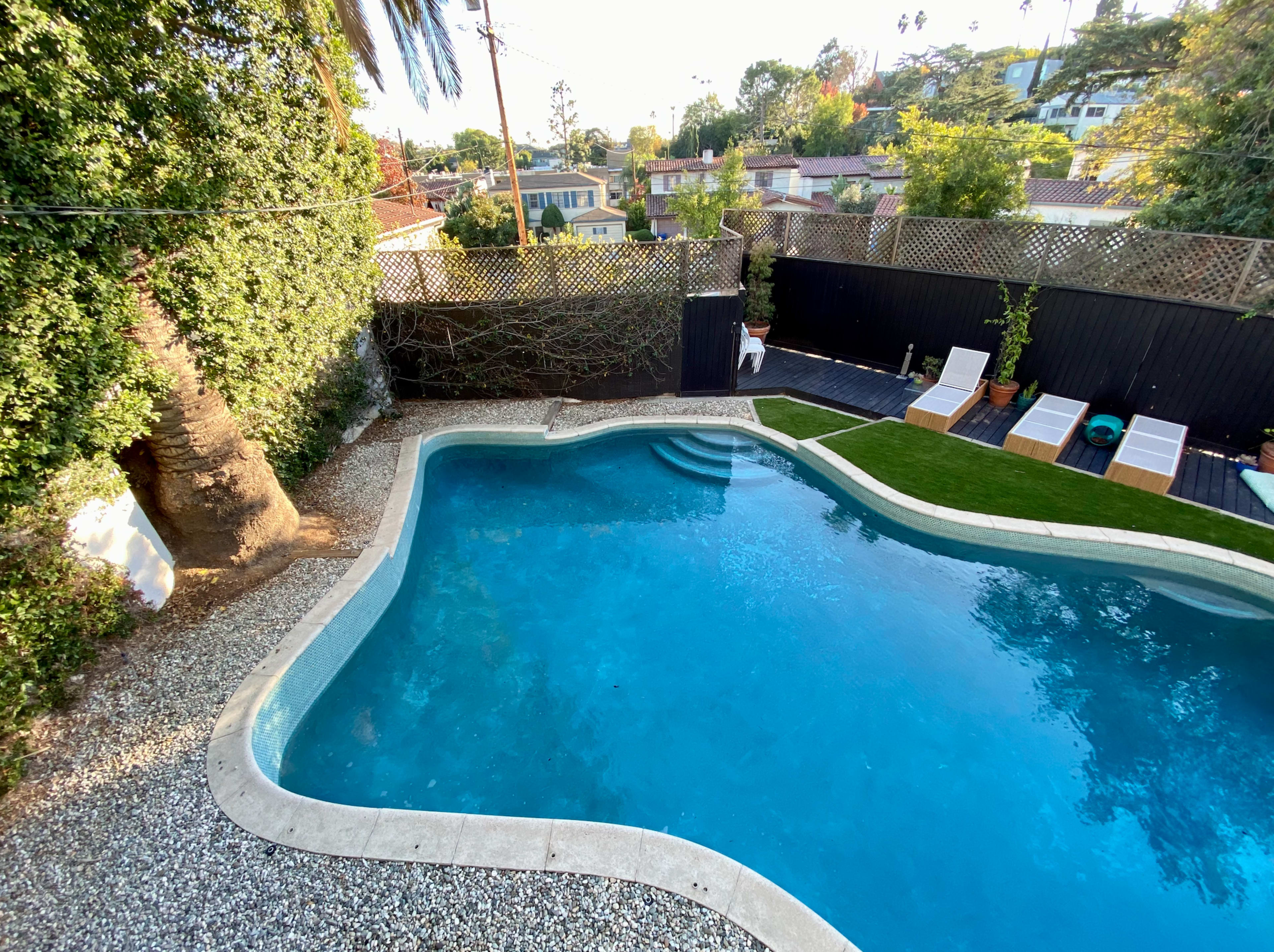 A swimming pool is surrounded by a gravel area and lounge chairs, with greenery and houses visible in the background.
