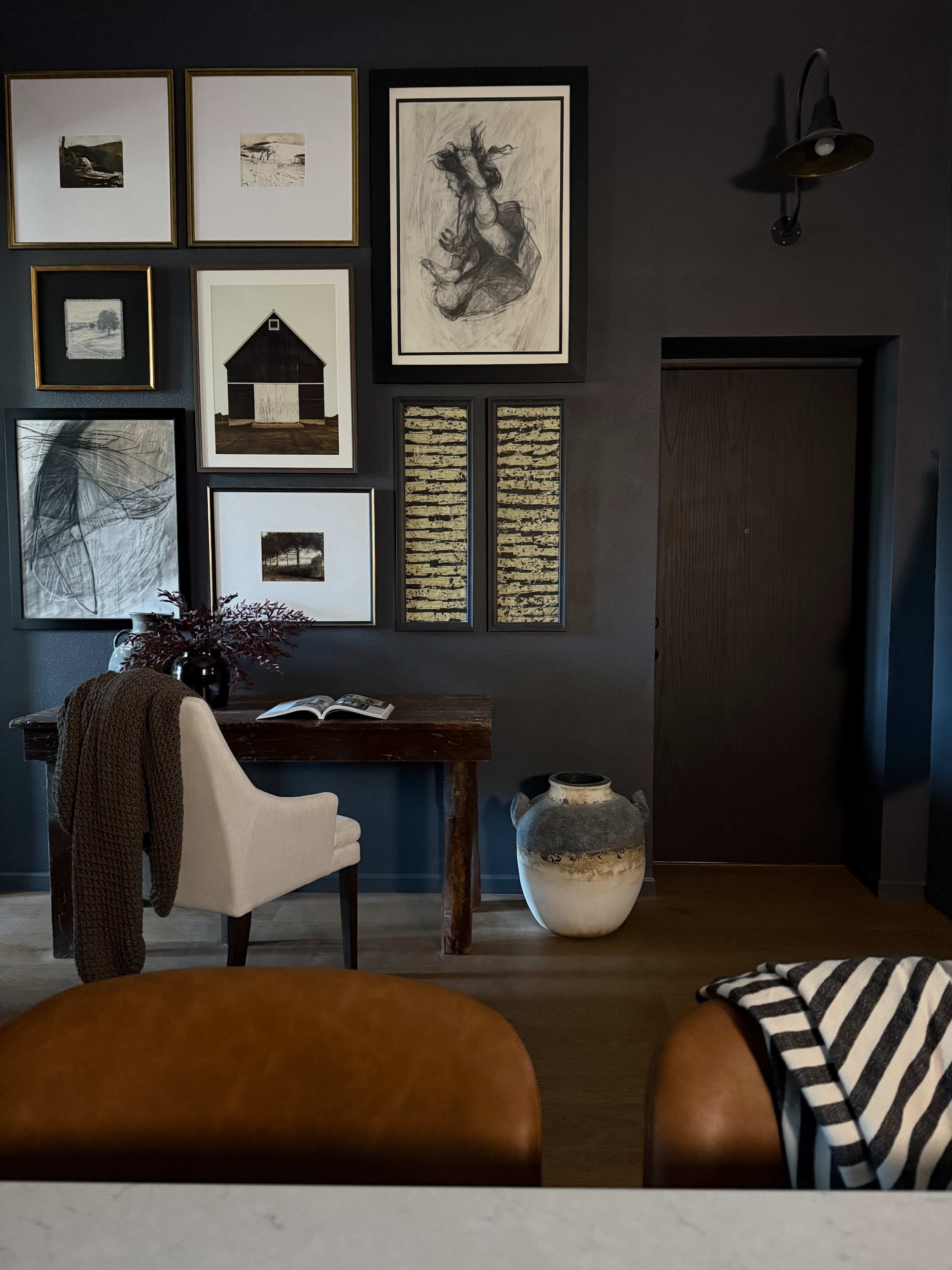 The image shows a dark room featuring a wooden table, a white chair, framed artwork on the wall, and a decorative vase beside an entrance door.