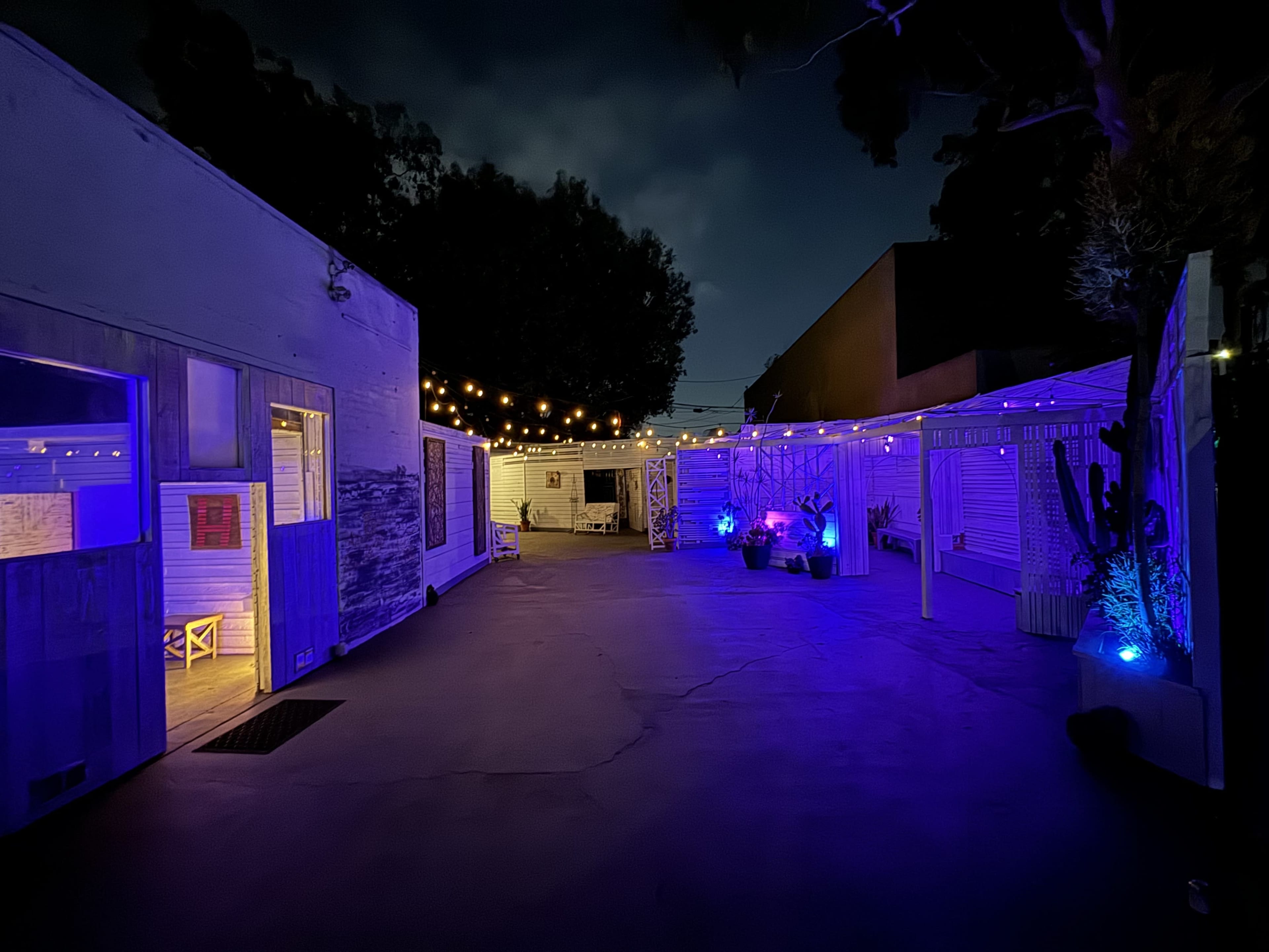 A dimly lit outdoor courtyard features string lights and purple illumination, surrounded by small buildings and potted plants.