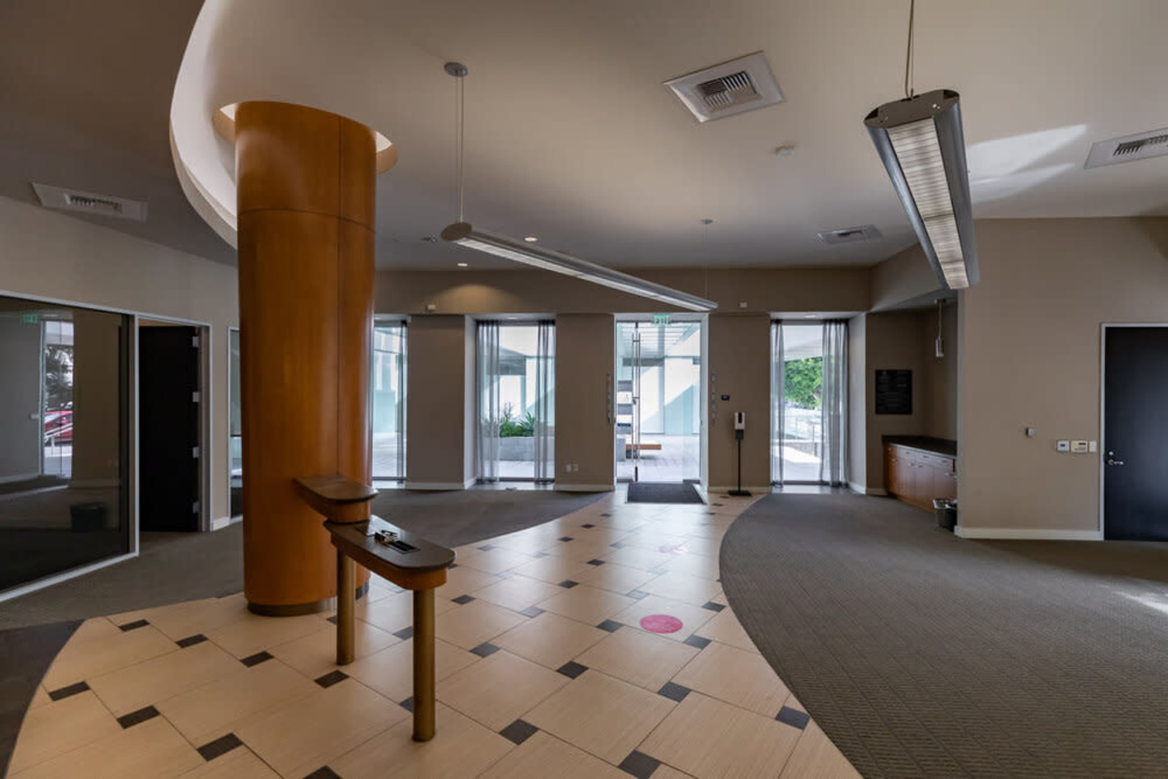 A modern lobby area with a circular wooden column, large glass doors, and a tiled floor featuring a geometric pattern.