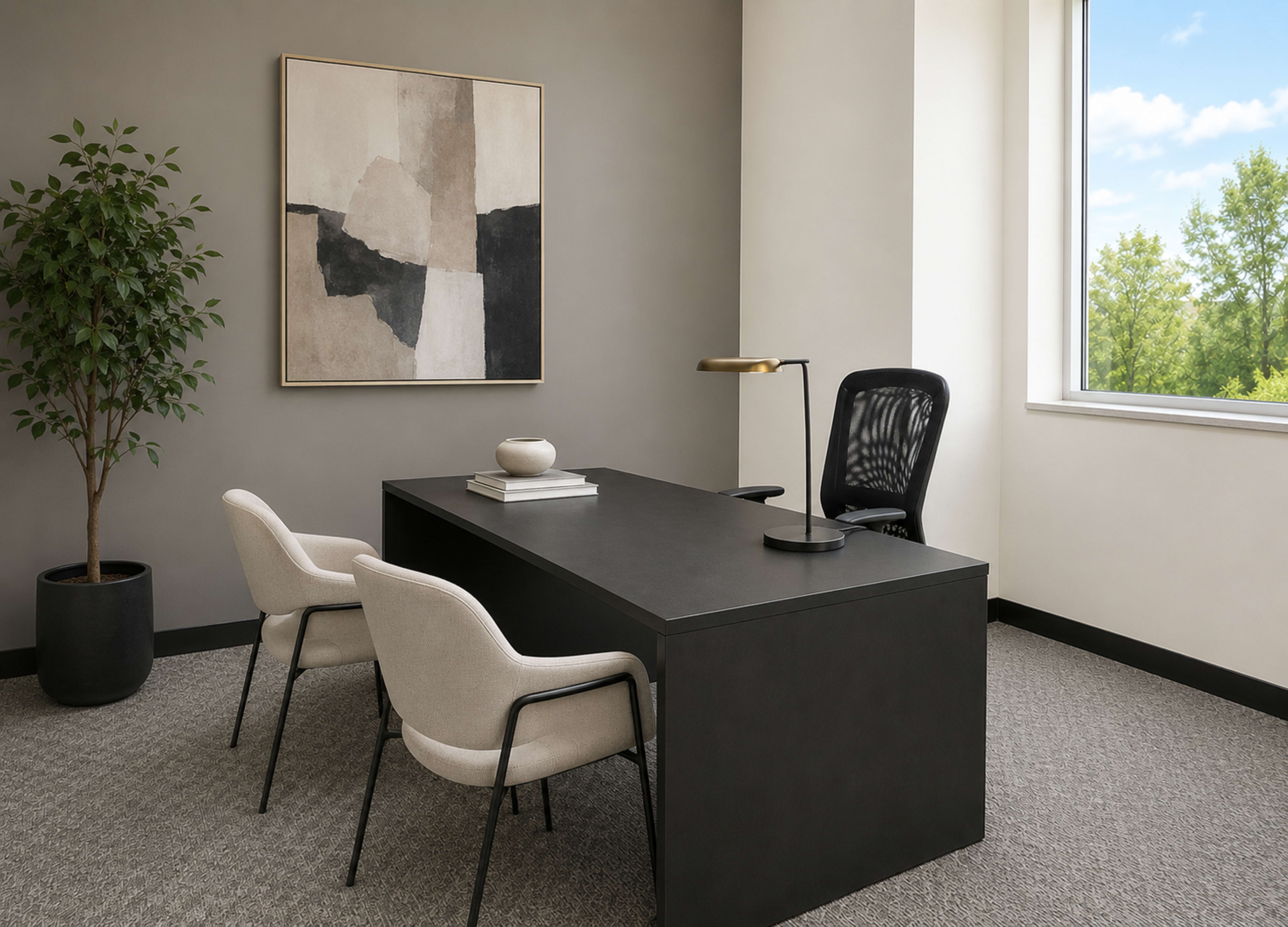 The image shows a modern office space with a large black desk, two cream-colored chairs, and a wall art piece, illuminated by natural light from a window overlooking greenery.