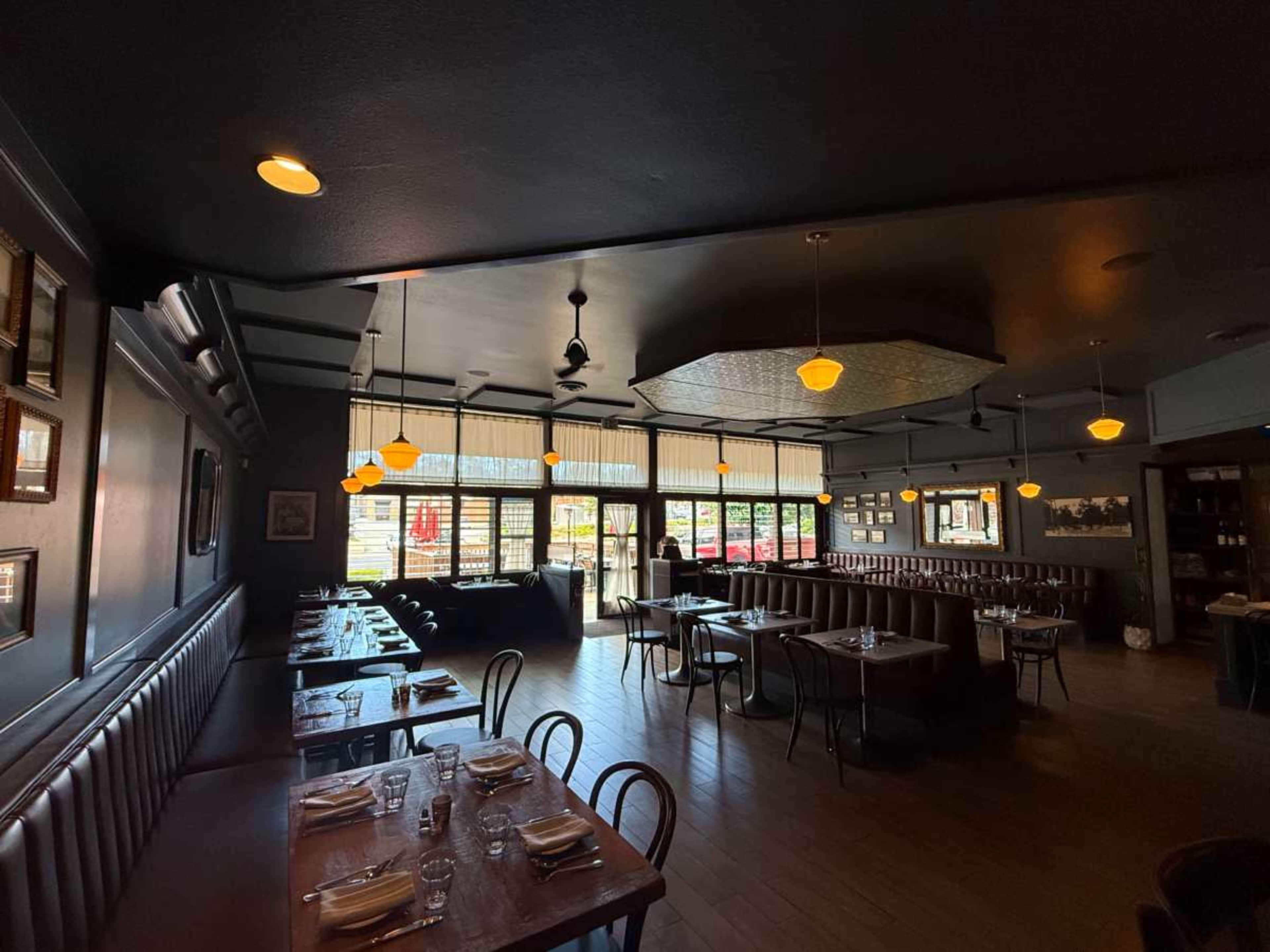 The image shows a dimly lit restaurant featuring wooden tables, bench seating, and large windows that open to an outdoor area.