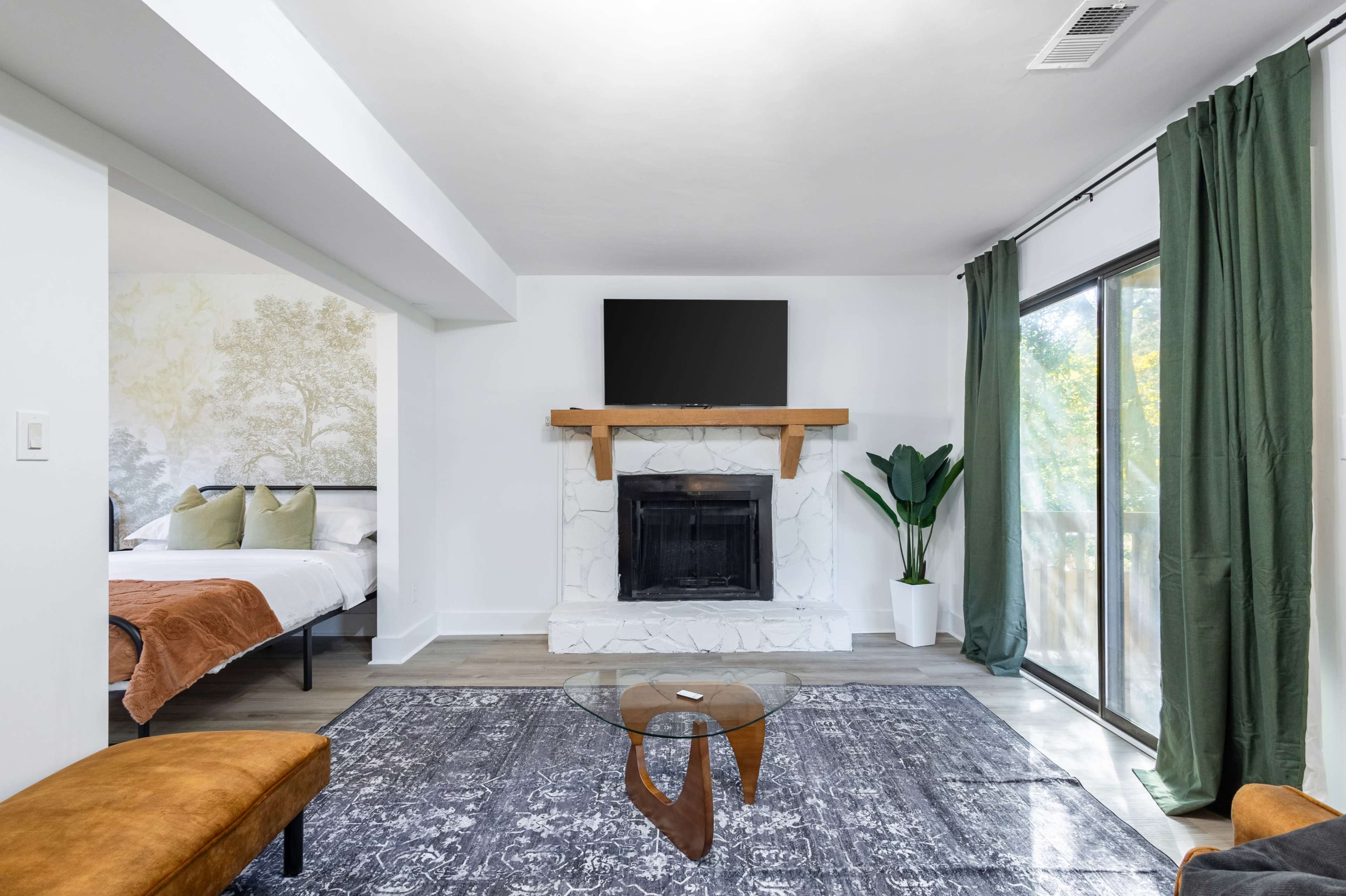 The image shows a modern living room with a fireplace, a mounted television, a glass coffee table, and large windows with green curtains.