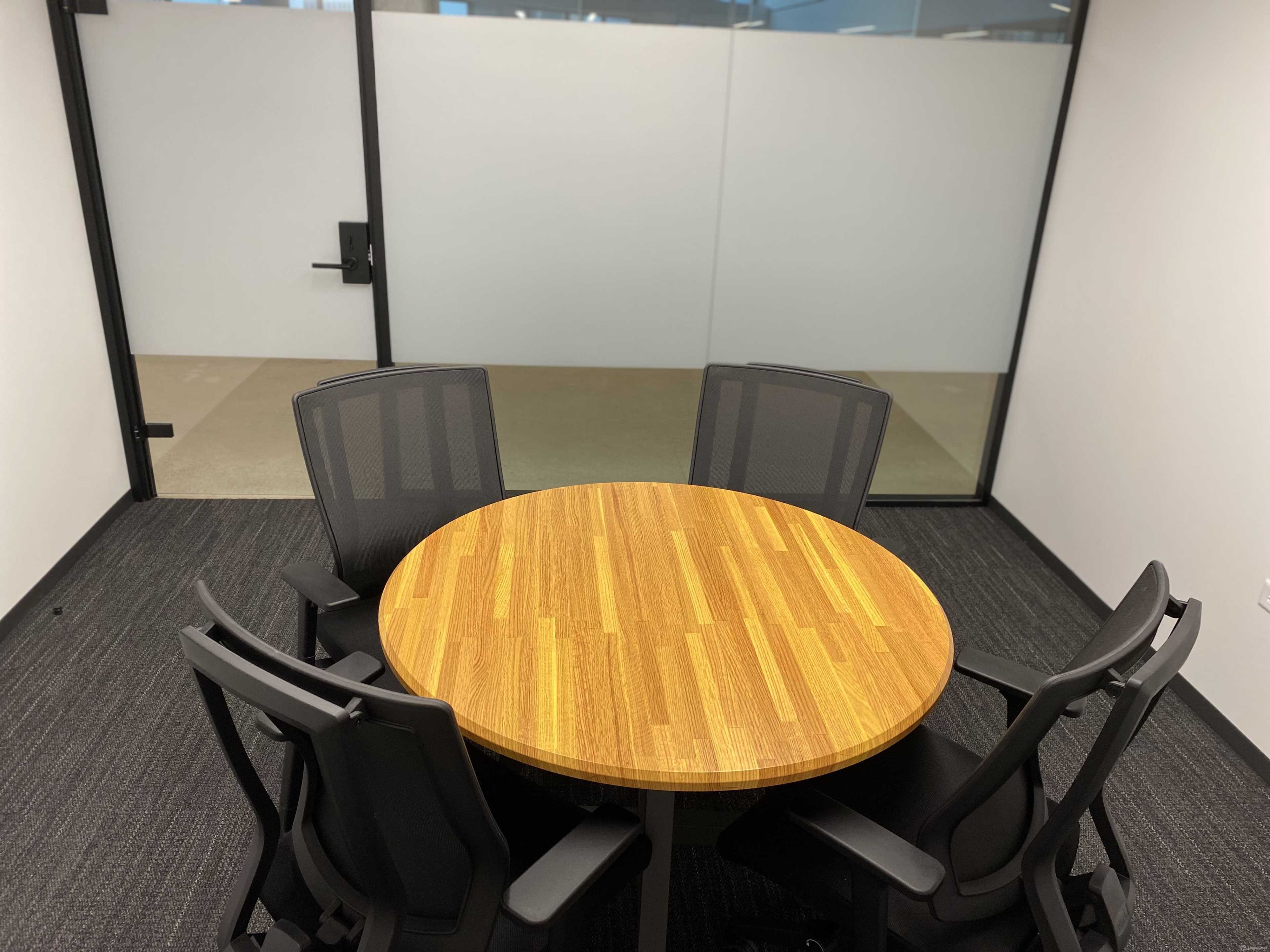 A round wooden table surrounded by four black mesh chairs in an office meeting room.