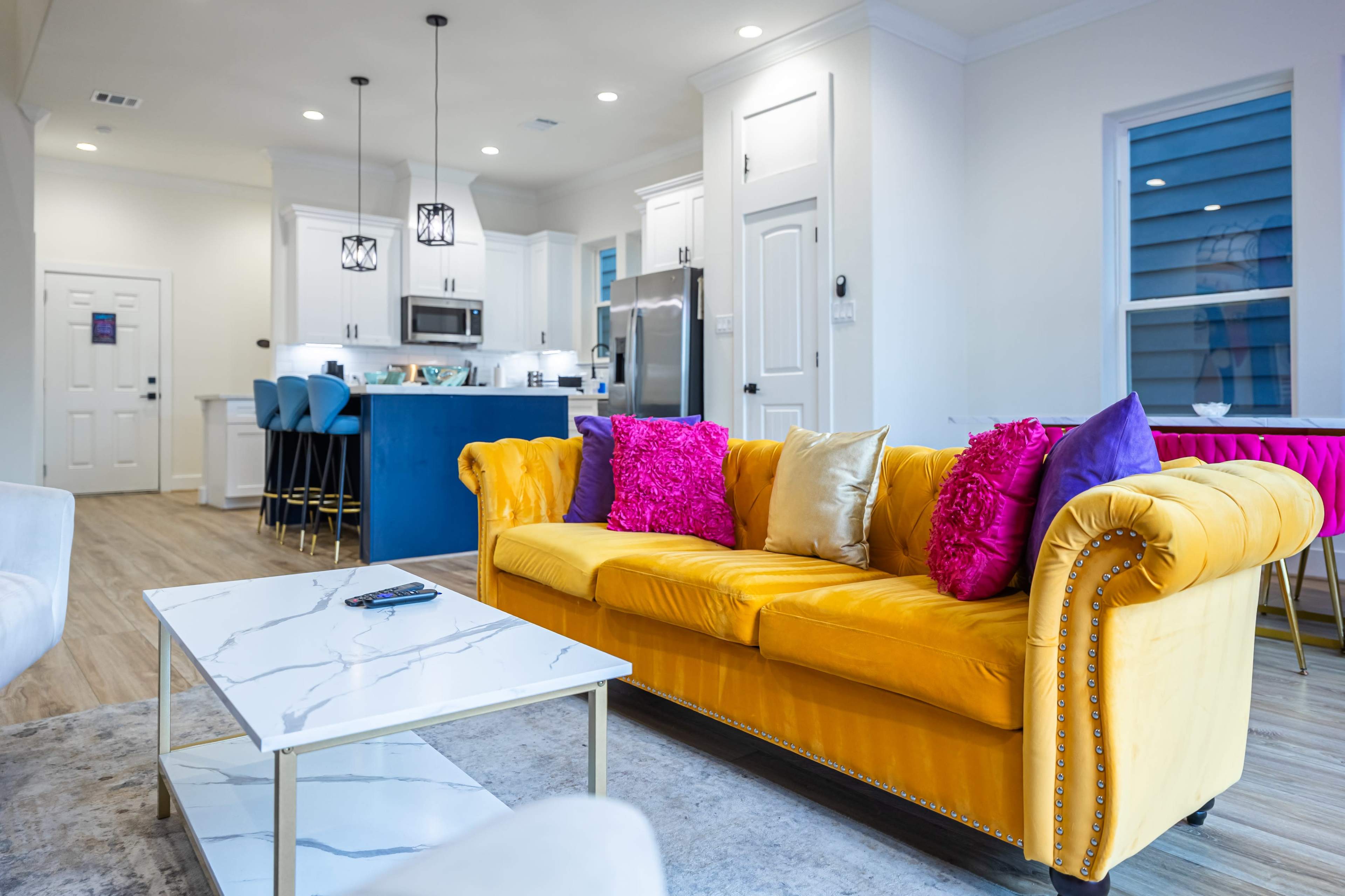 A bright yellow sofa with colorful cushions is positioned in a modern living room that leads to a kitchen area with white cabinetry and bar seating.