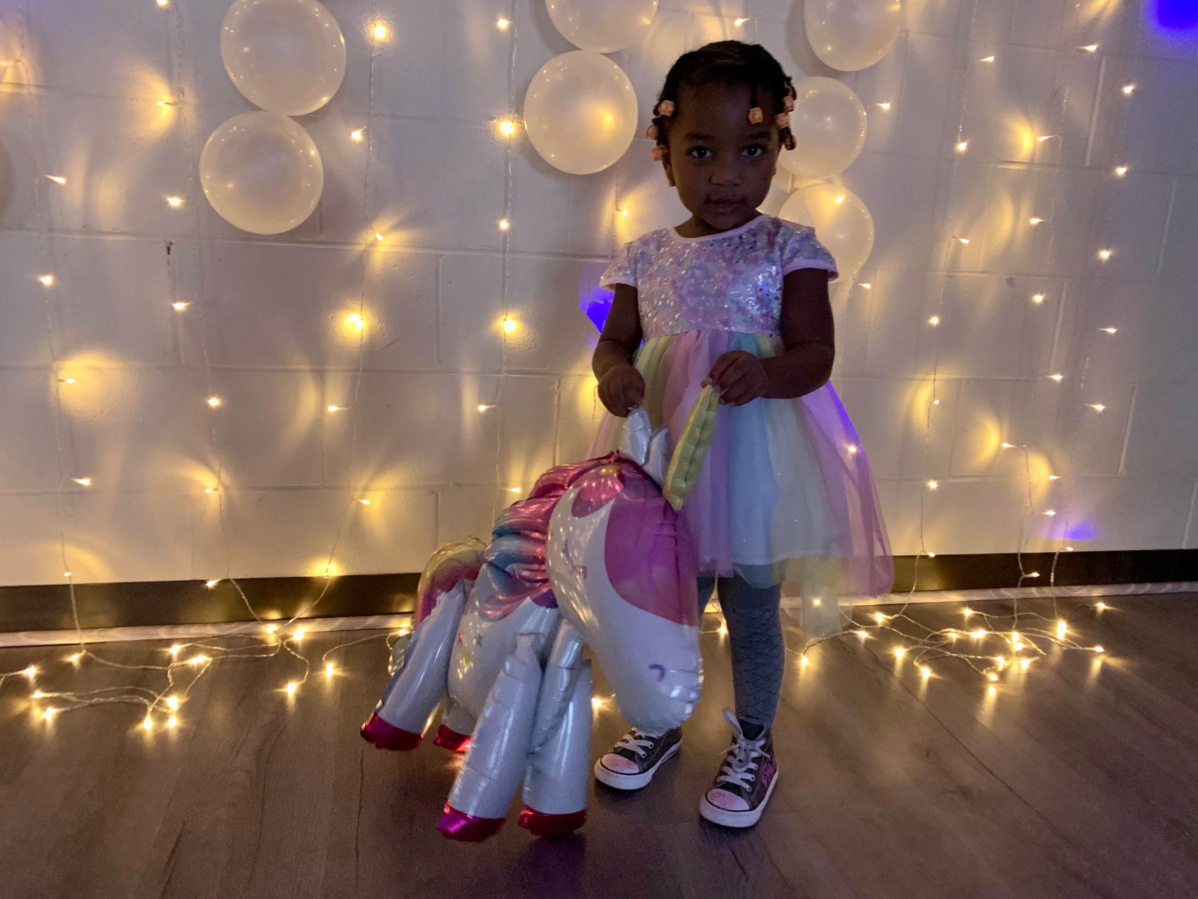 A young girl in a colorful dress holds a unicorn-shaped inflatable toy against a backdrop of soft glowing lights.