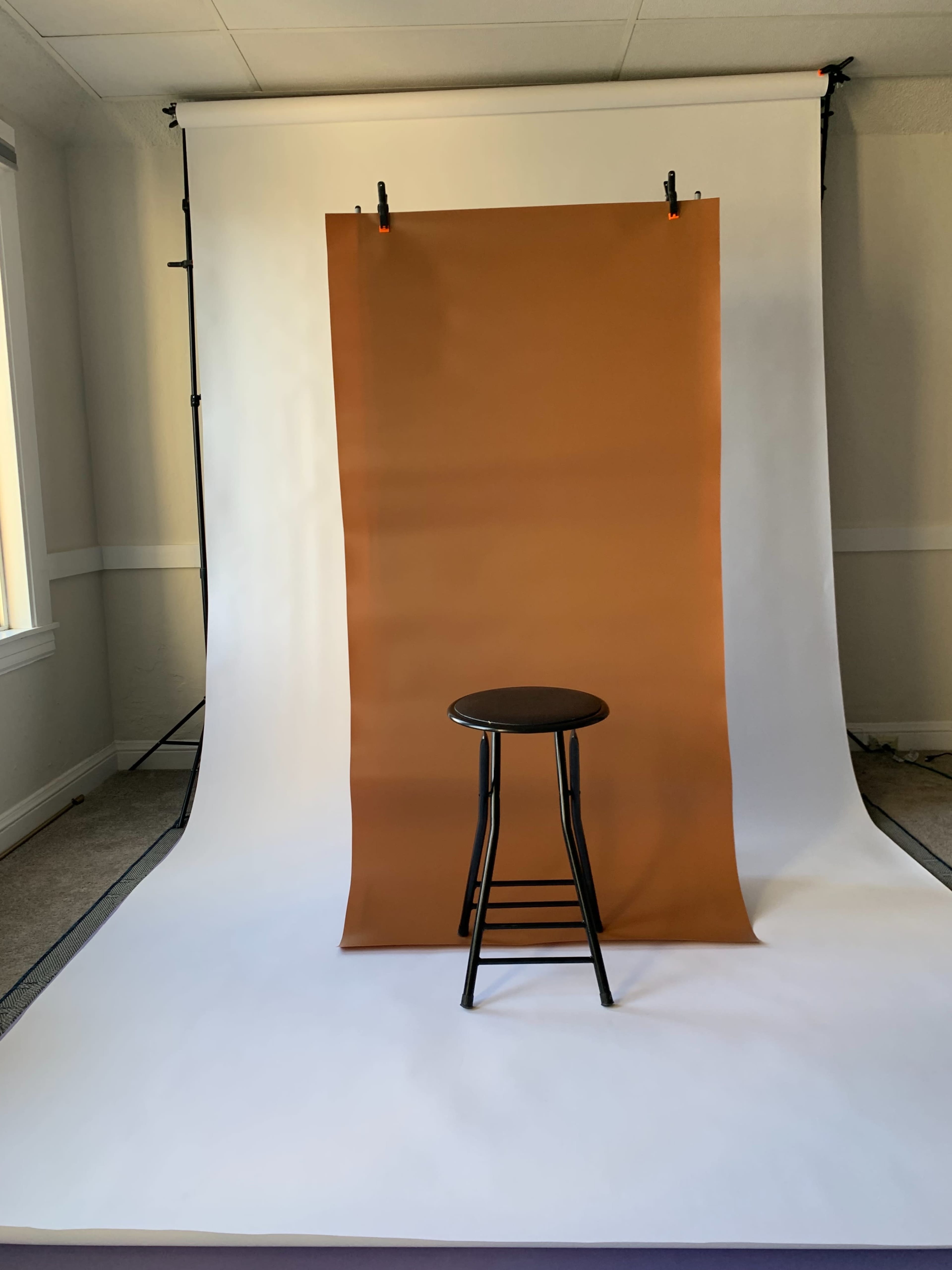 A black stool is positioned in front of a brown backdrop suspended from a stand against a white backdrop.