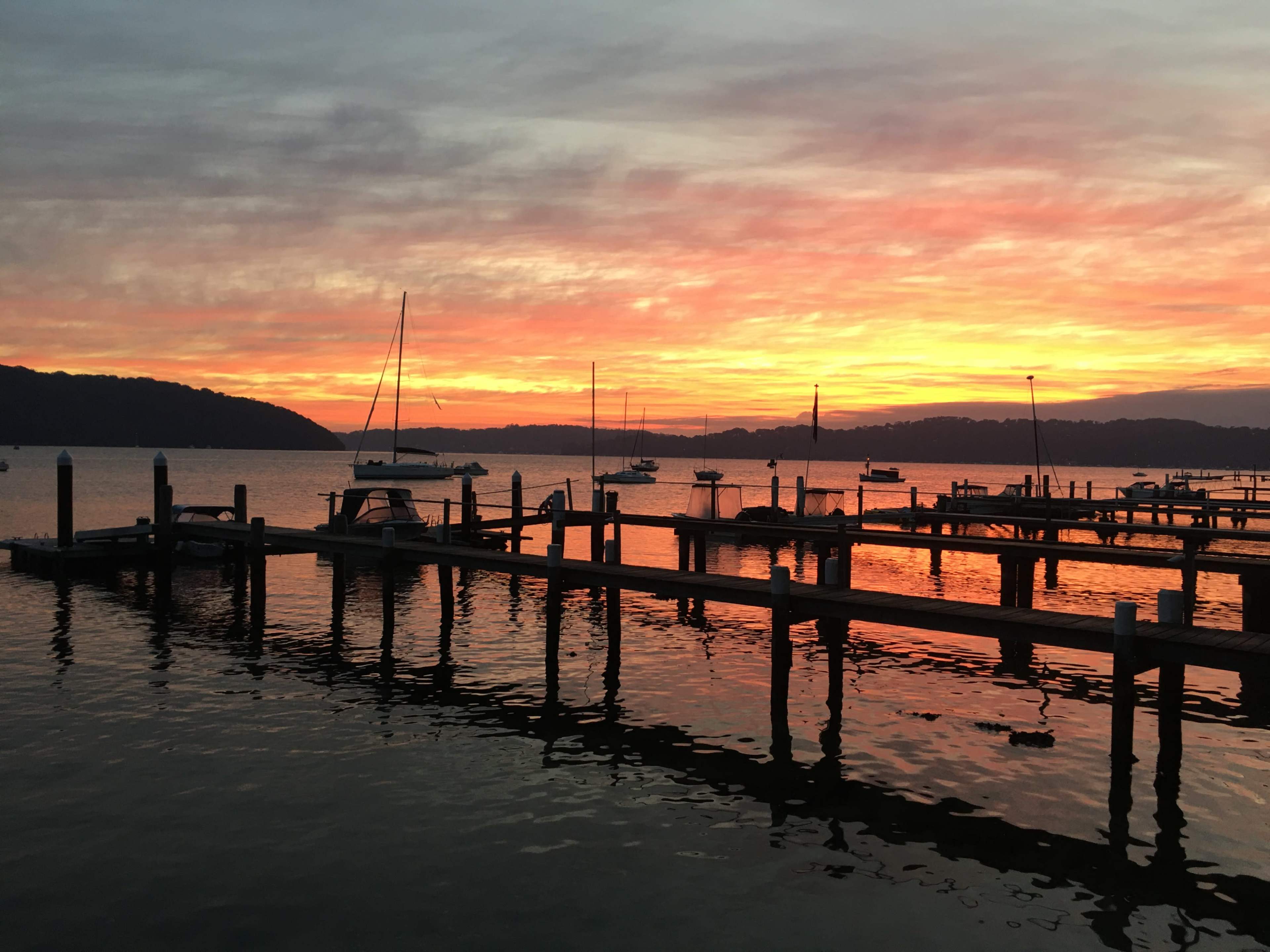 The image shows a marina at sunset, with boats moored along wooden docks and a colorful sky reflected in the water.
