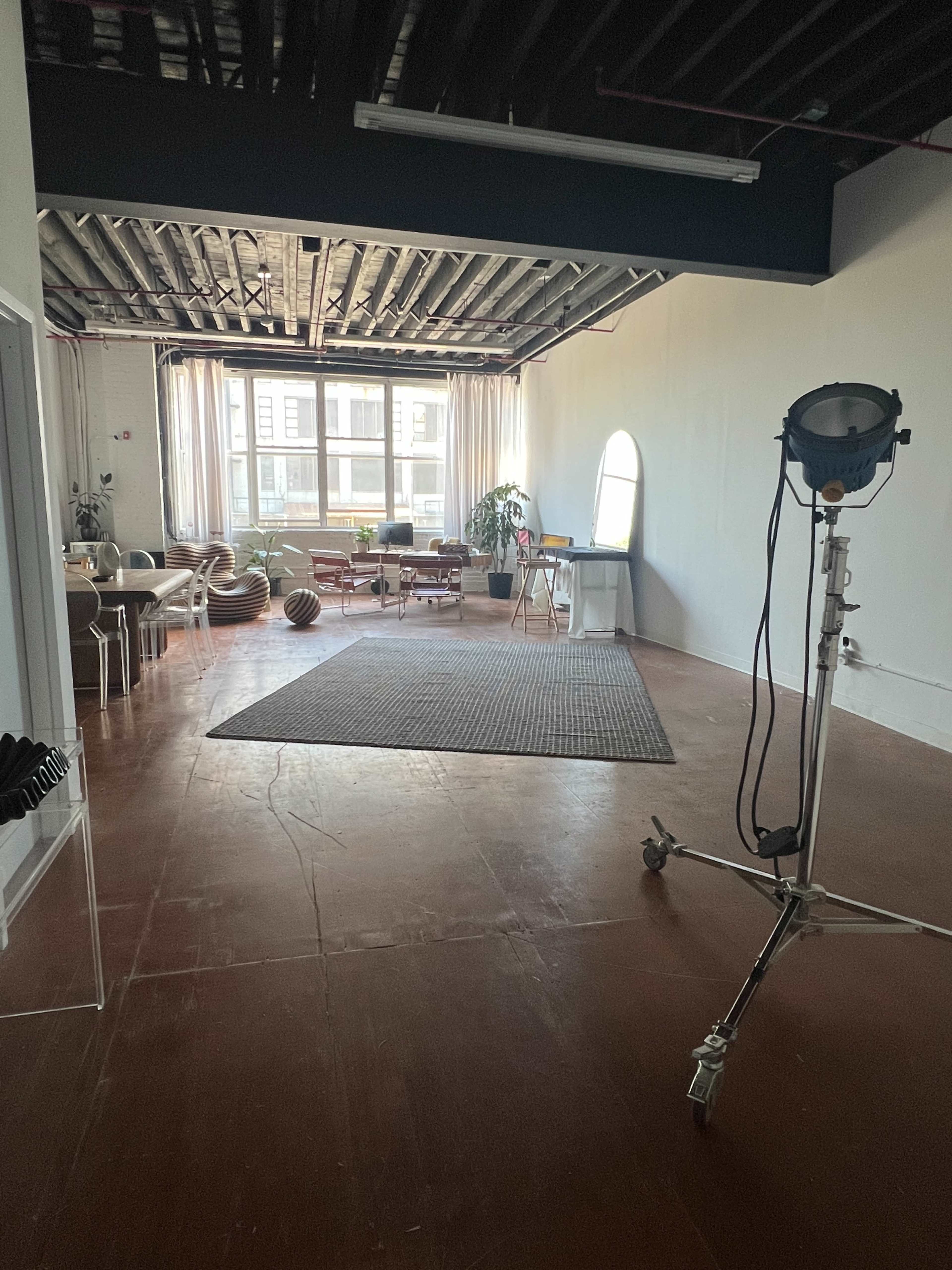 The image shows a spacious interior room with large windows, featuring a seating area, a rug on the floor, and a lighting setup on a movable stand.