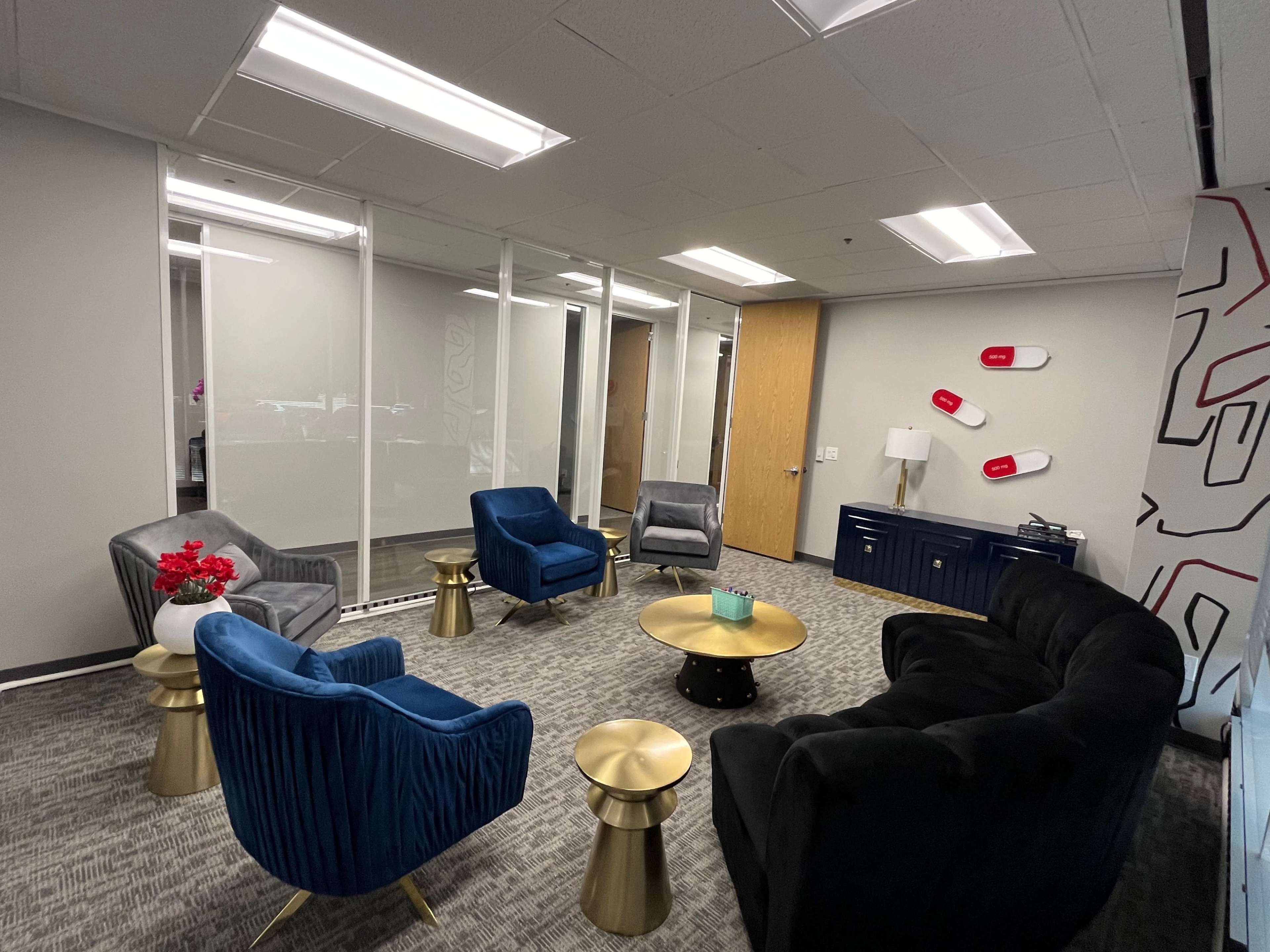 The image shows a modern office lounge area with a mix of upholstered seating, a small brass coffee table, and decorative elements against a neutral wall.