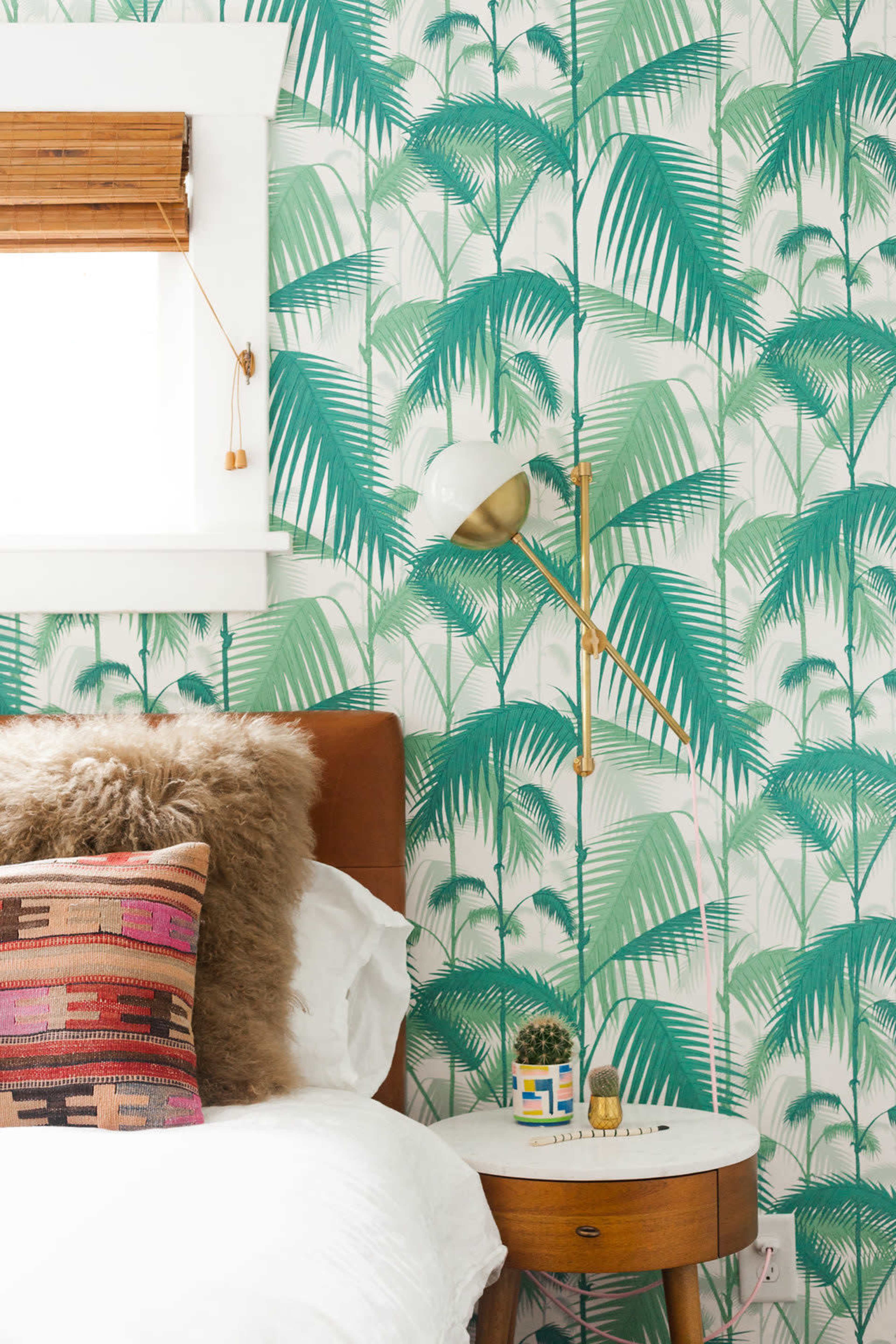 The image shows a well-decorated bedroom featuring a palm leaf-patterned wallpaper, a brown leather bed, a white bedspread, and a side table with a lamp and decorative items.