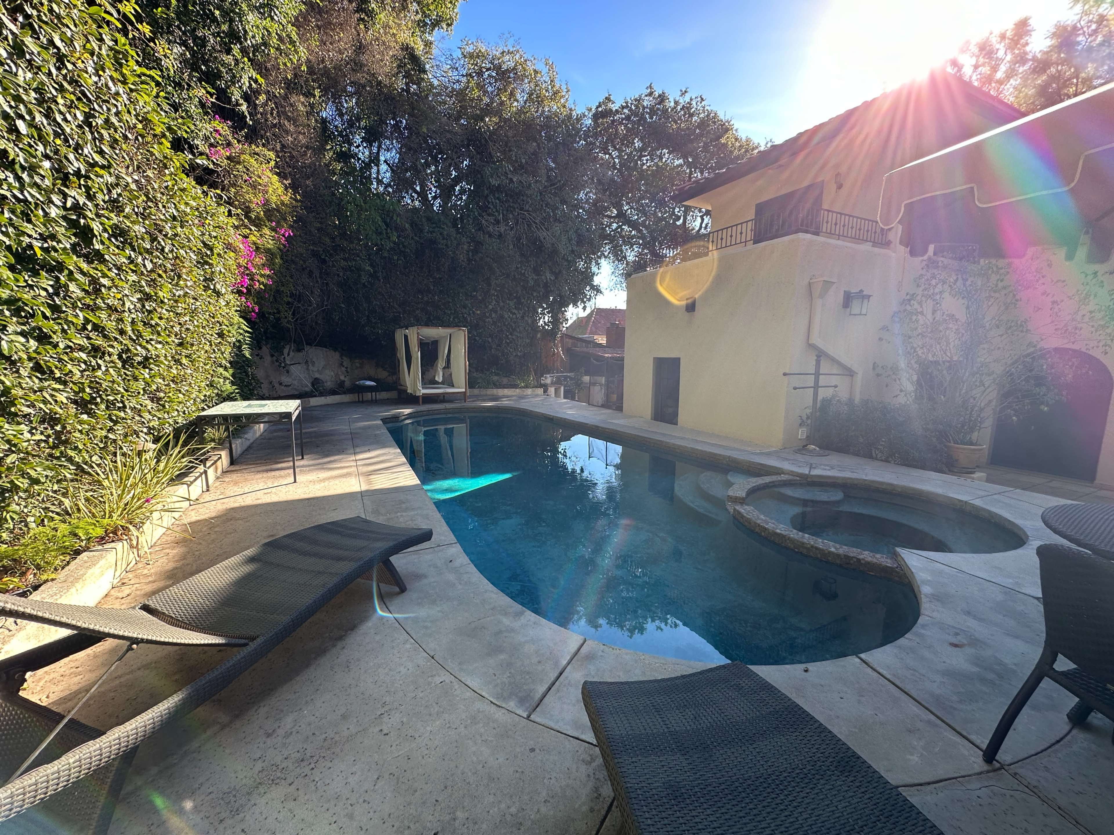 The image shows a residential backyard with a swimming pool, a spa, and lounge chairs surrounded by lush greenery and a yellow house partially in view.
