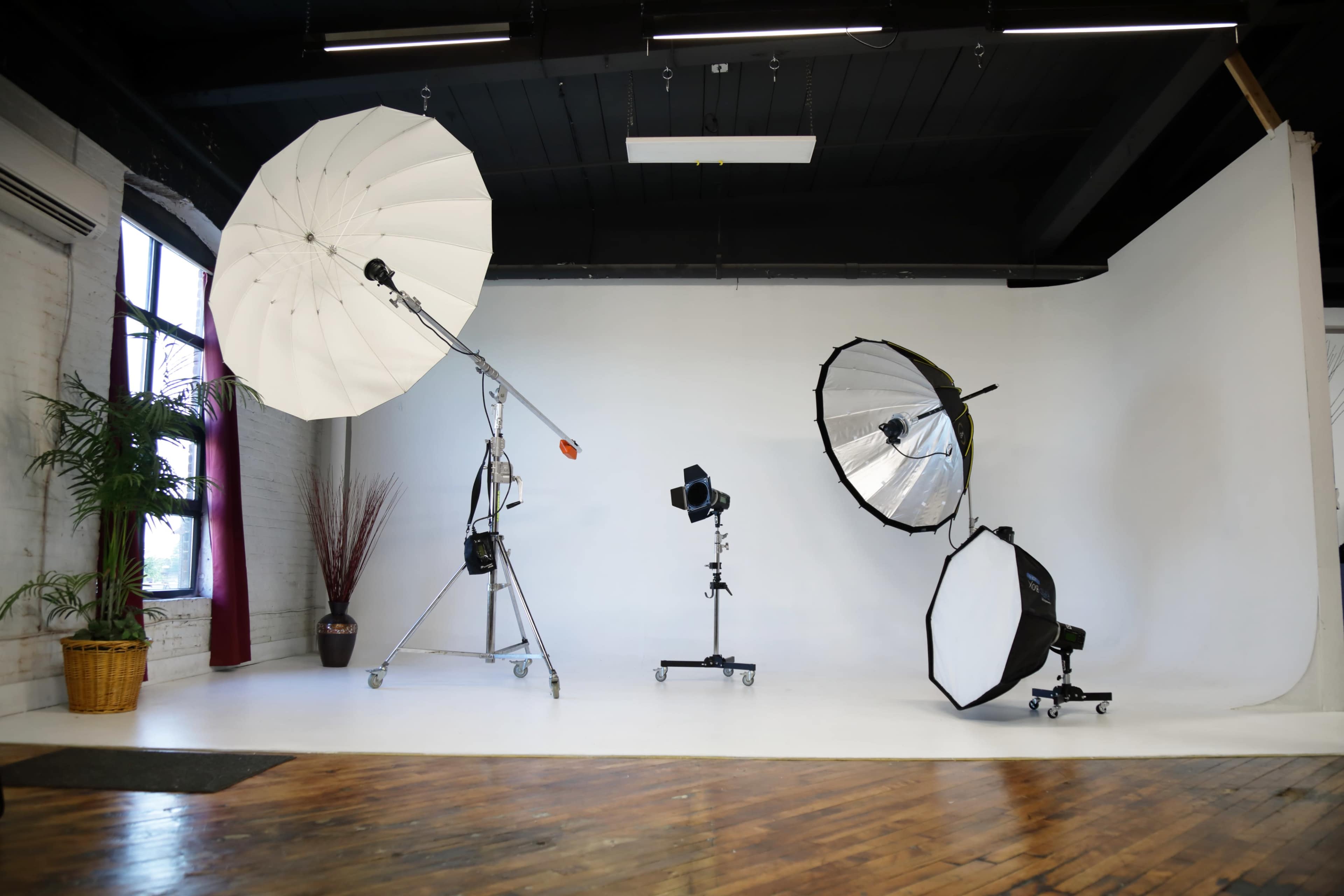 The image shows a photography studio with various lighting equipment, including a large umbrella, a beauty dish, and a softbox, arranged in front of a white backdrop.