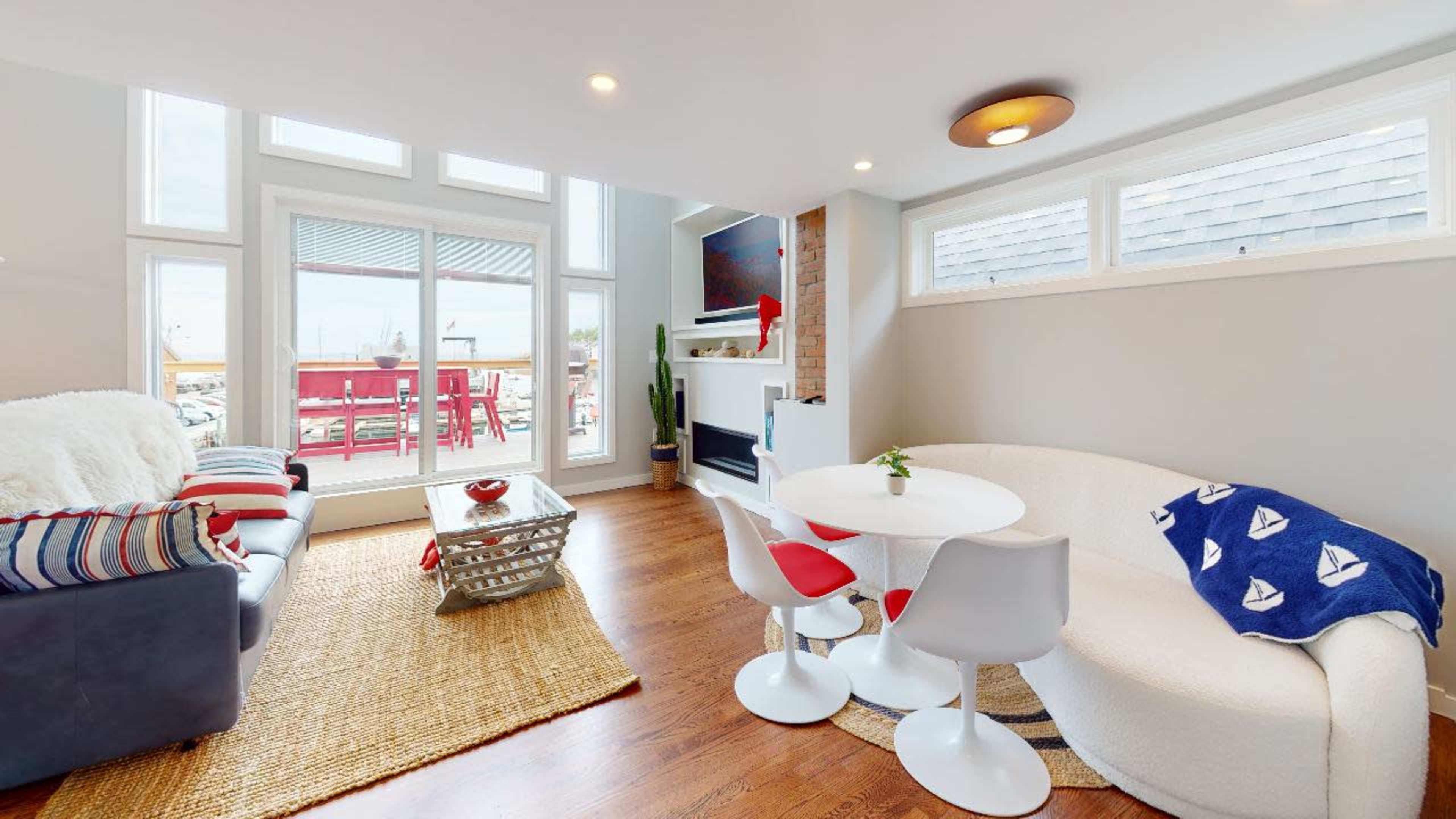 A modern living room features a light-colored sofa, a round dining table with red and white chairs, and large windows overlooking a patio.