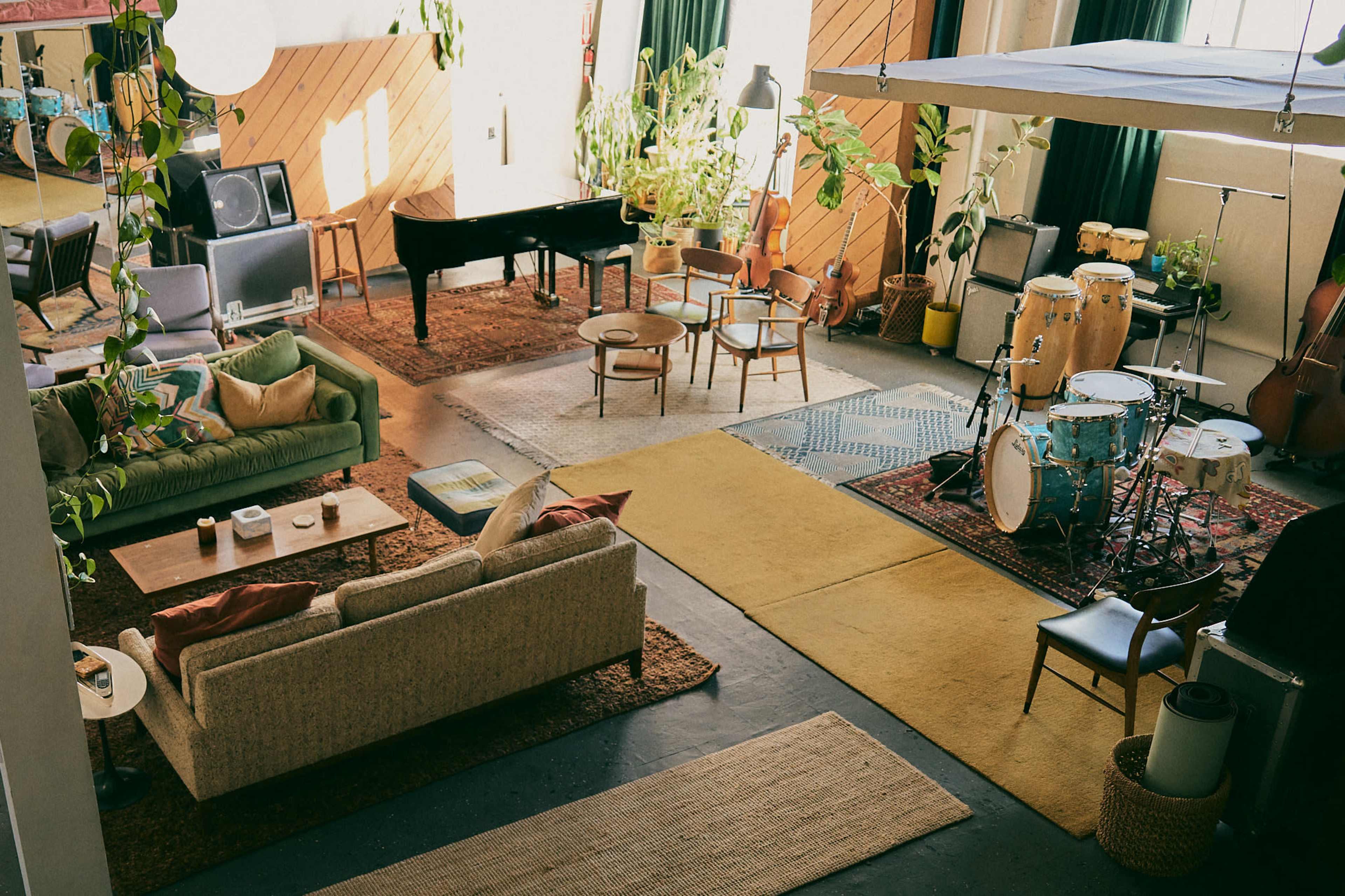 The image shows a spacious, well-decorated room with a piano, seating areas, plants, and musical instruments, arranged on various rugs.