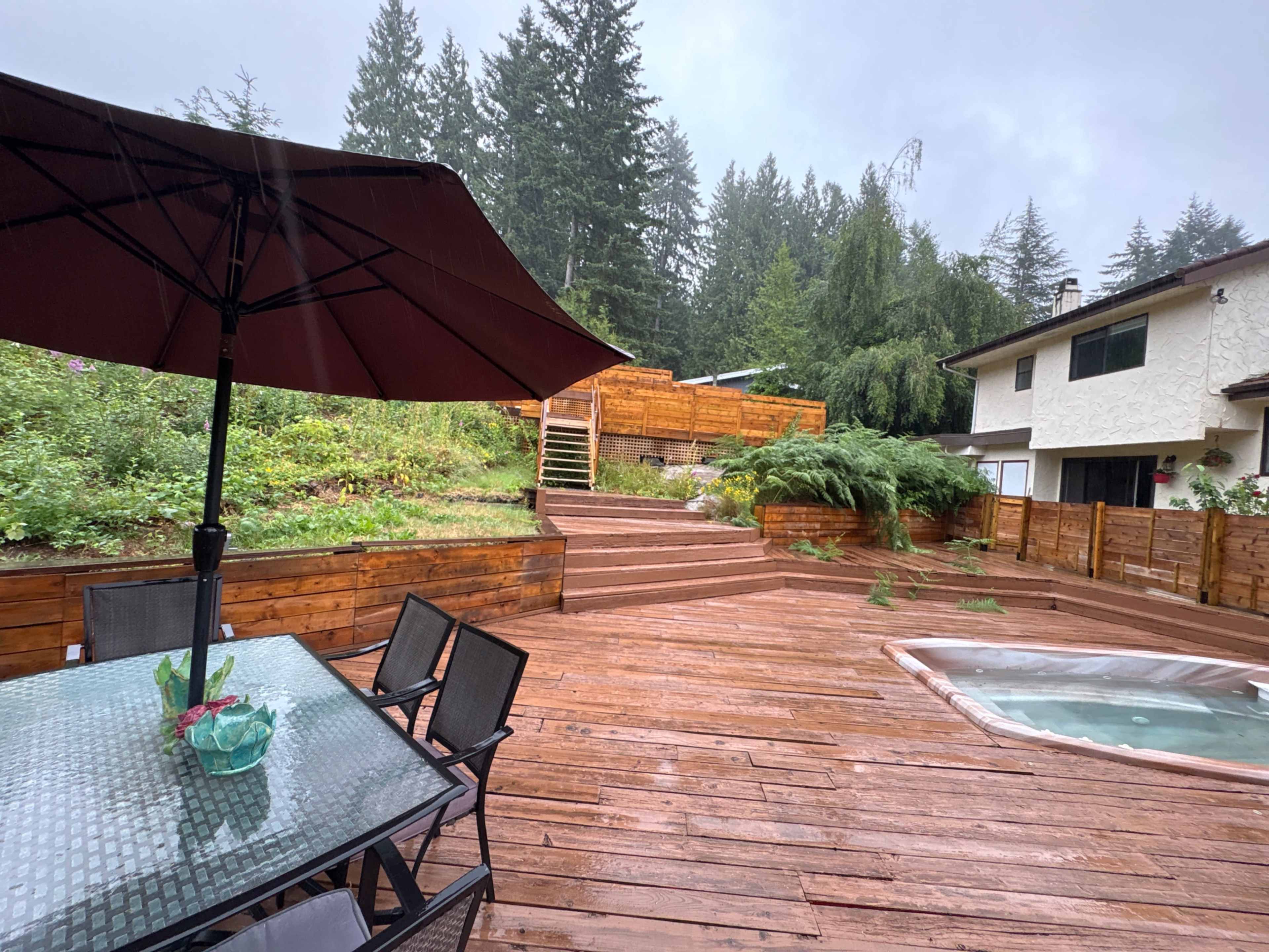 The image shows a wooden deck area with patio furniture, a hot tub, and an uphill garden surrounded by trees on a rainy day.