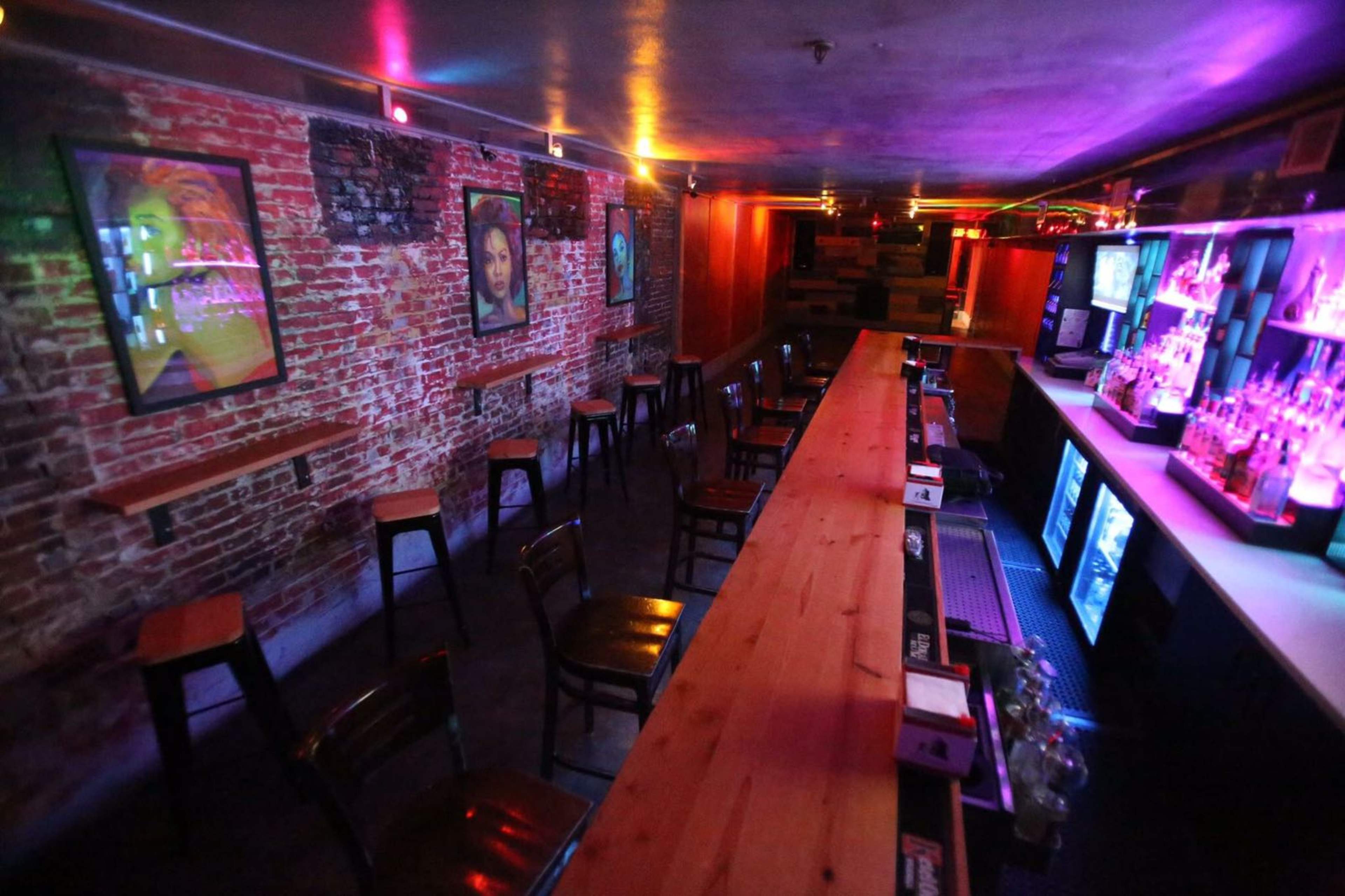 An empty bar area featuring a long wooden counter, high-backed chairs, exposed brick walls with framed artwork, and colorful lighting.