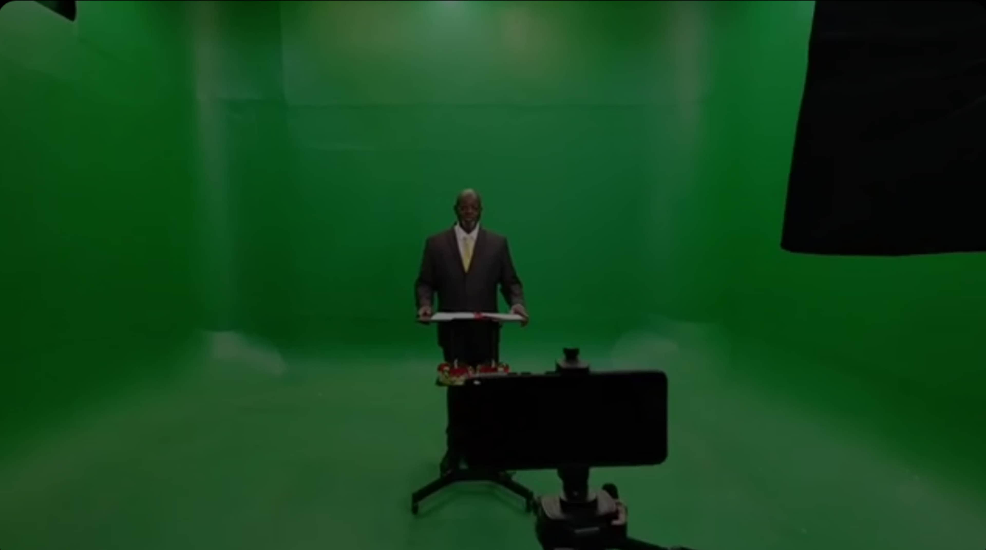 A speaker stands at a podium in front of a green screen setup.