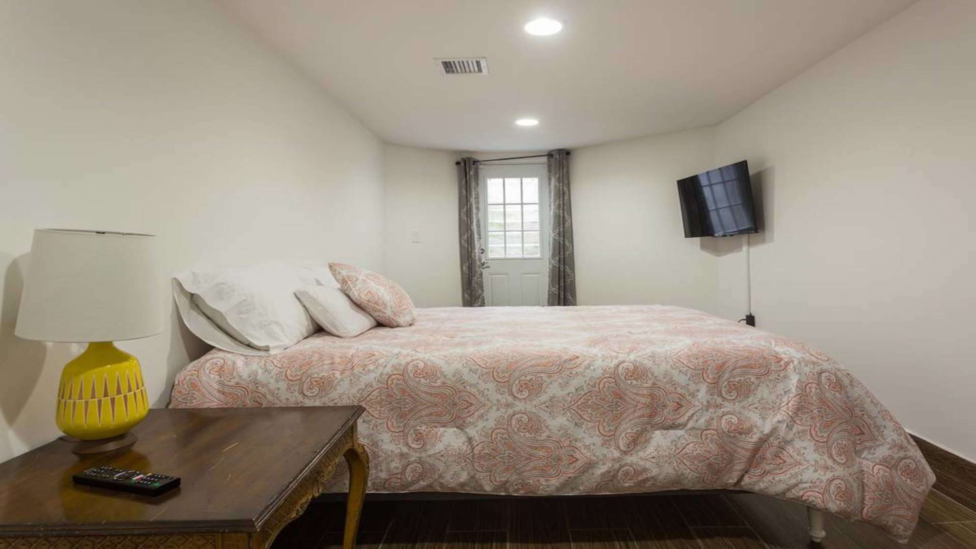 The image shows a small bedroom with a bed, a table lamp, a TV mounted on the wall, and a window with curtains.