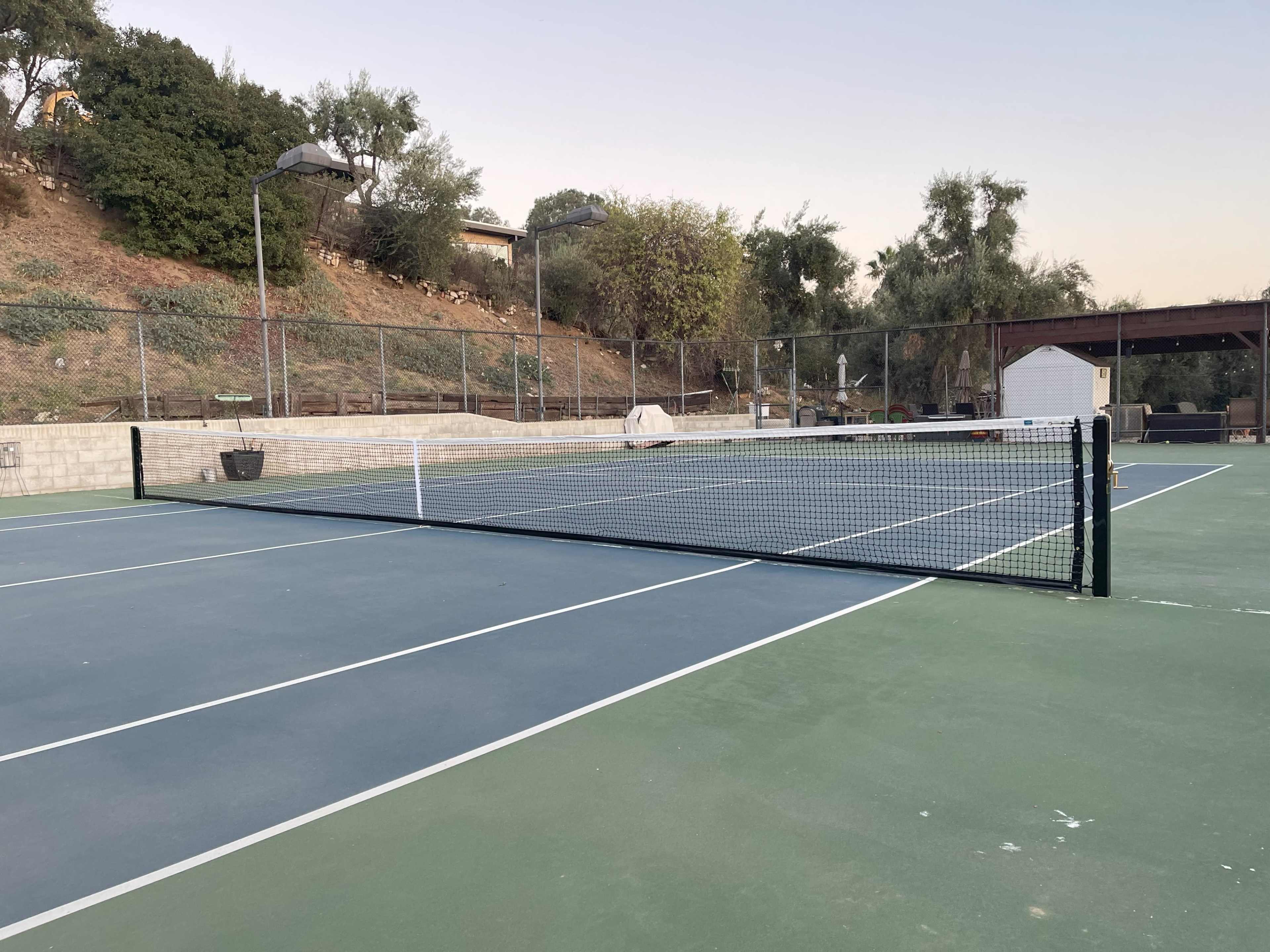 A tennis court with a net in the foreground, surrounded by trees and a small building in the background.