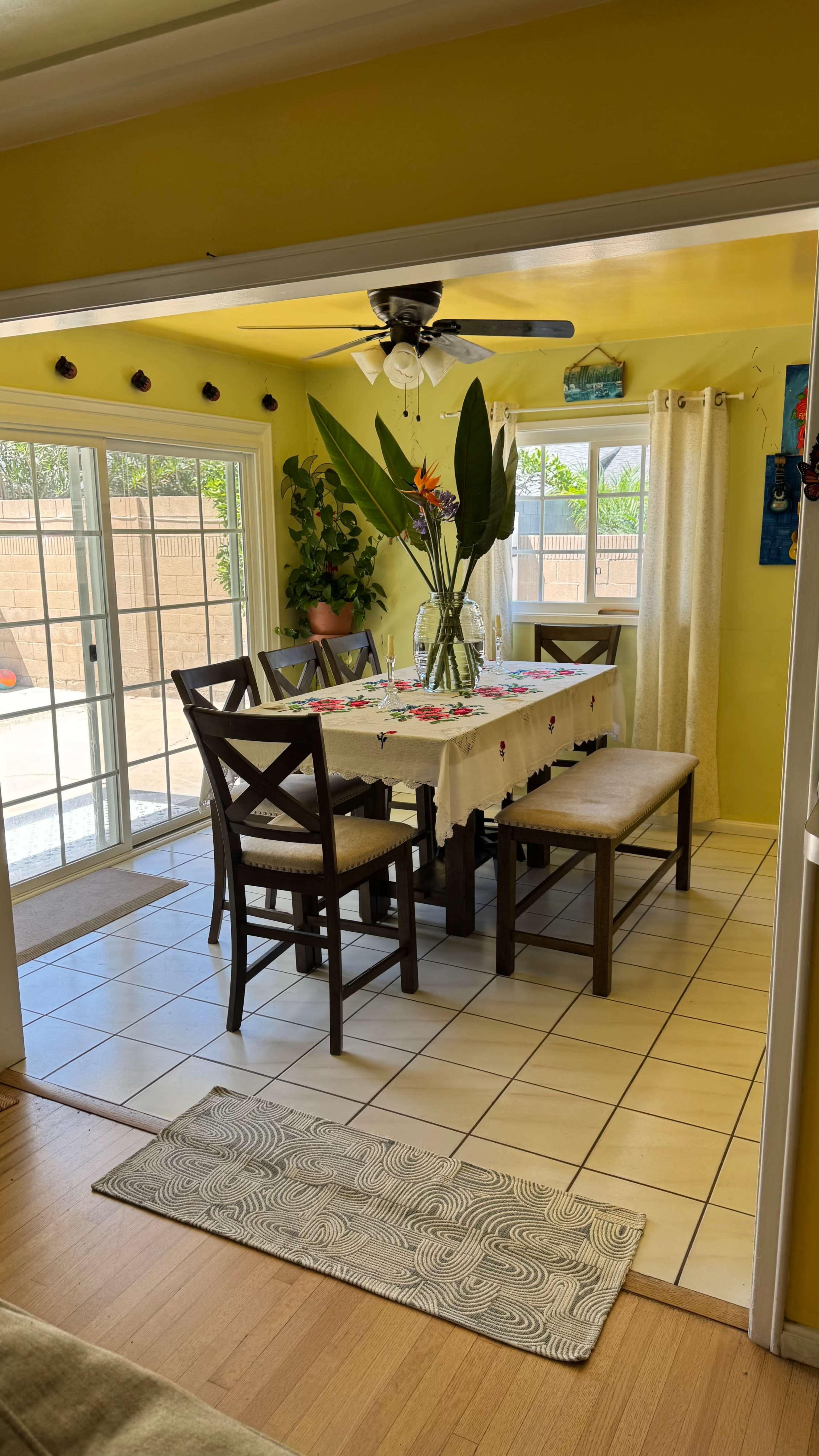 The image shows a bright yellow dining area featuring a large table set with a floral tablecloth, surrounded by chairs, with large windows letting in natural light.