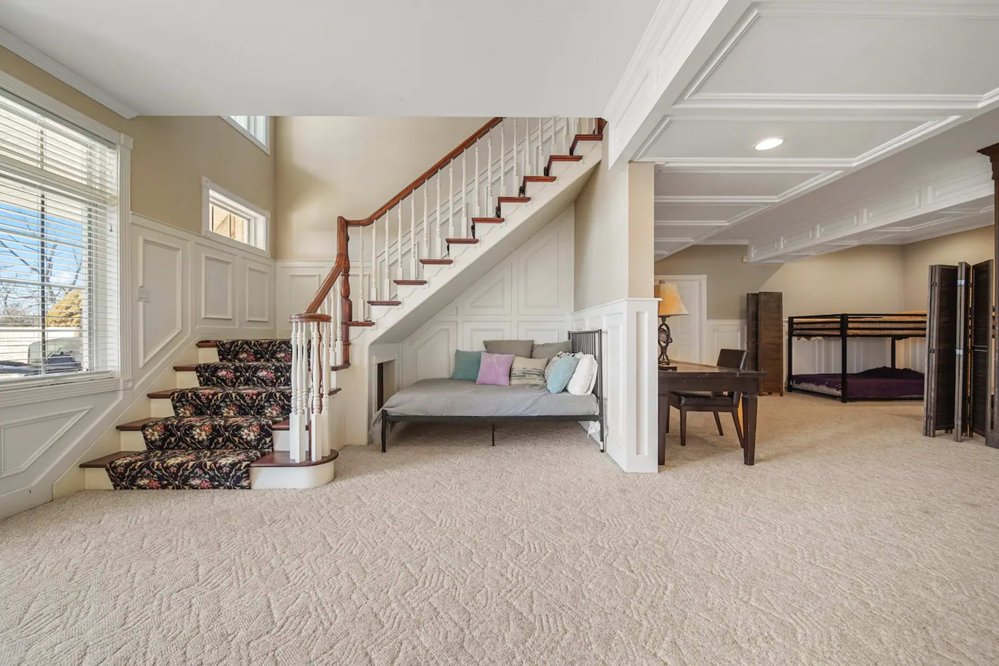 A spacious living area with a staircase, a seating area, a desk, and a bunk bed in a well-lit basement.