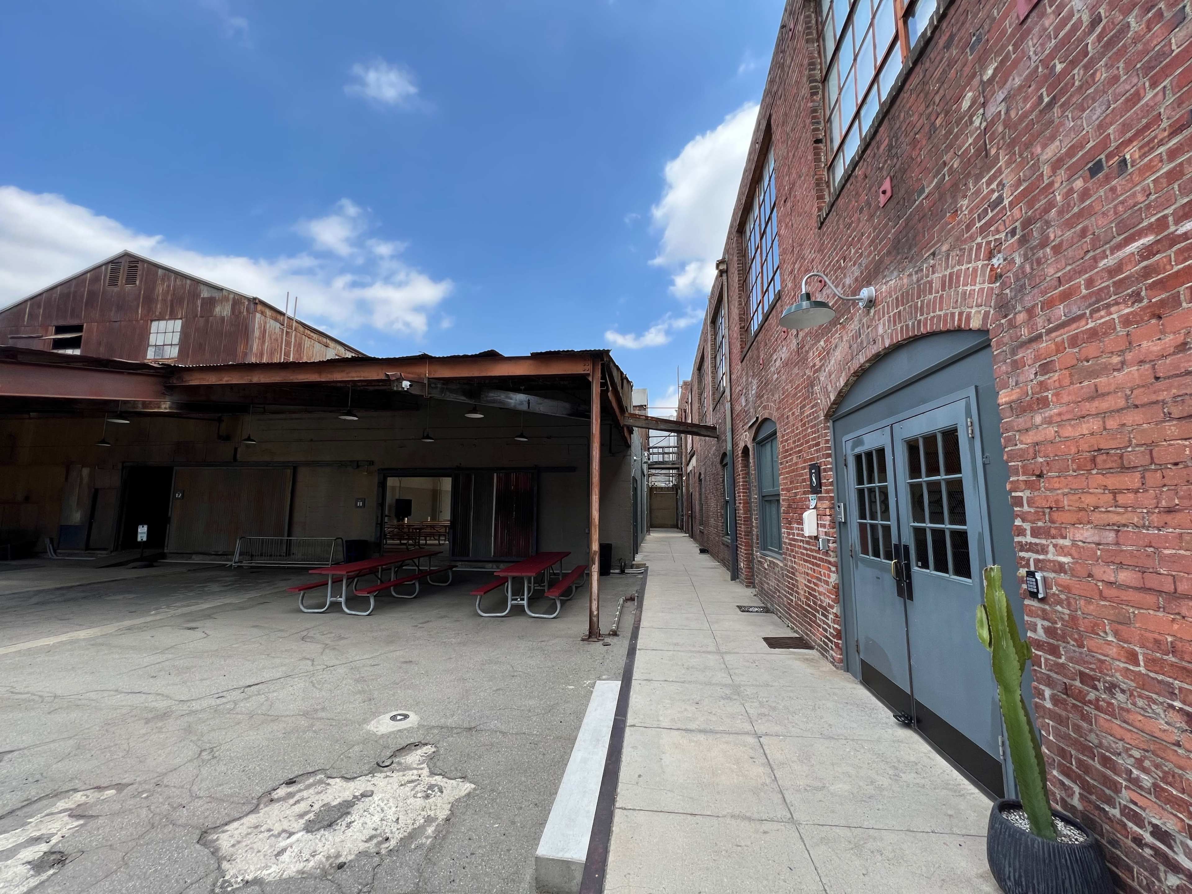 A narrow walkway beside a brick building with large windows, leading to a shaded outdoor area with picnic tables.