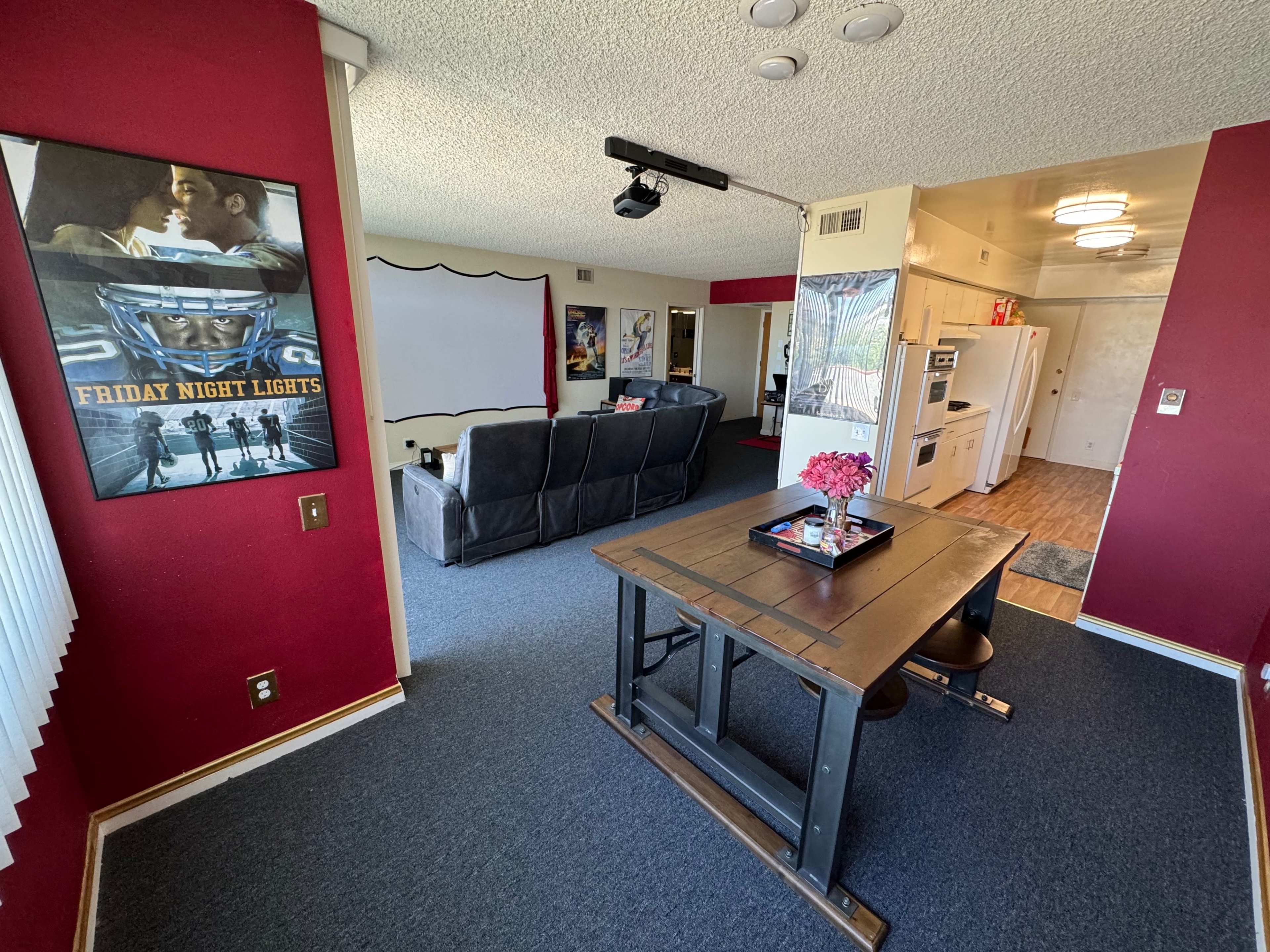A living space with a dining table at the foreground, a seating area with gray couches in the background, and a large blank wall for a projector or screen.