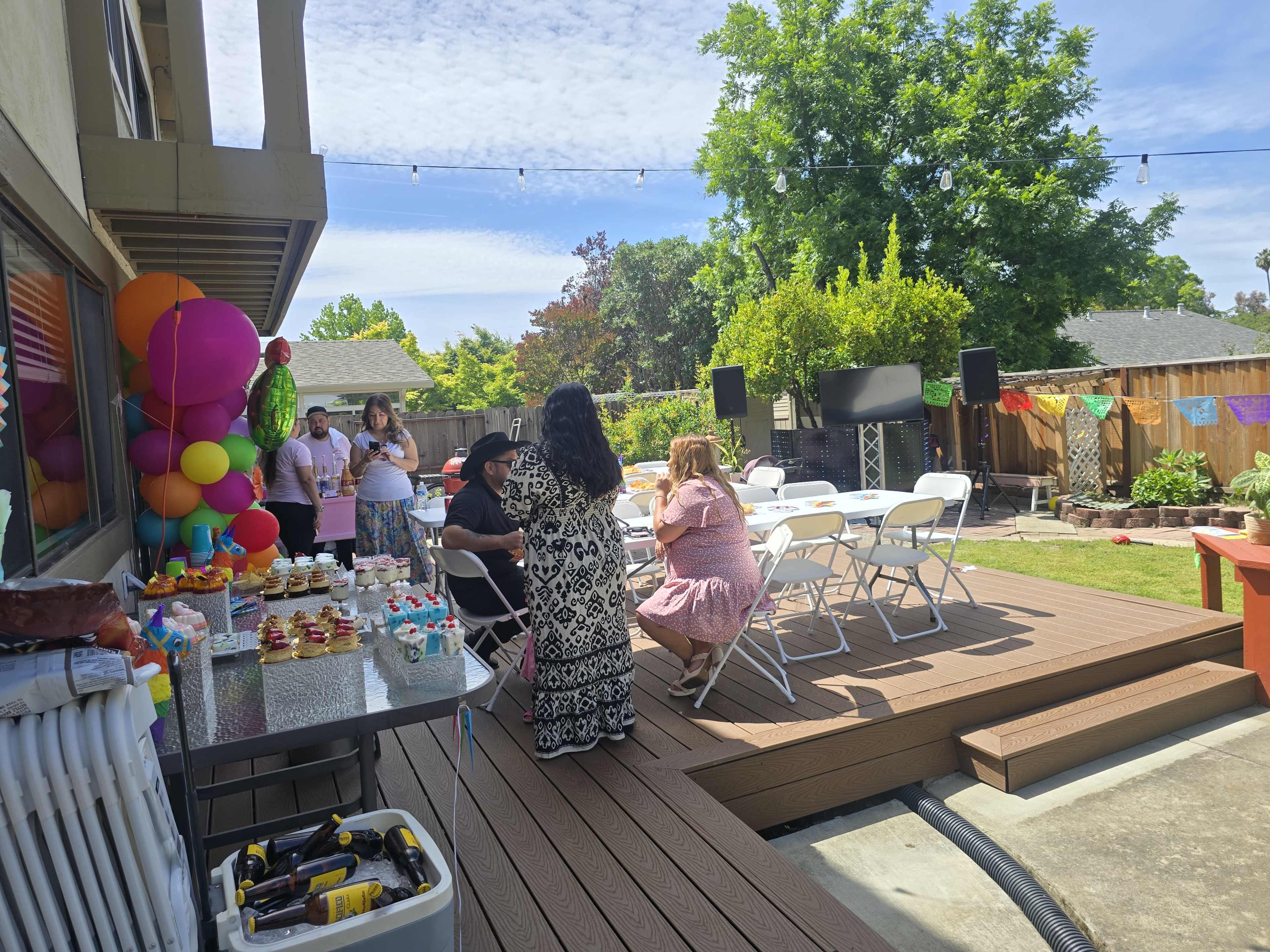 A group of people gathers in a backyard setting decorated with colorful balloons and a table filled with food and drinks for a celebration.
