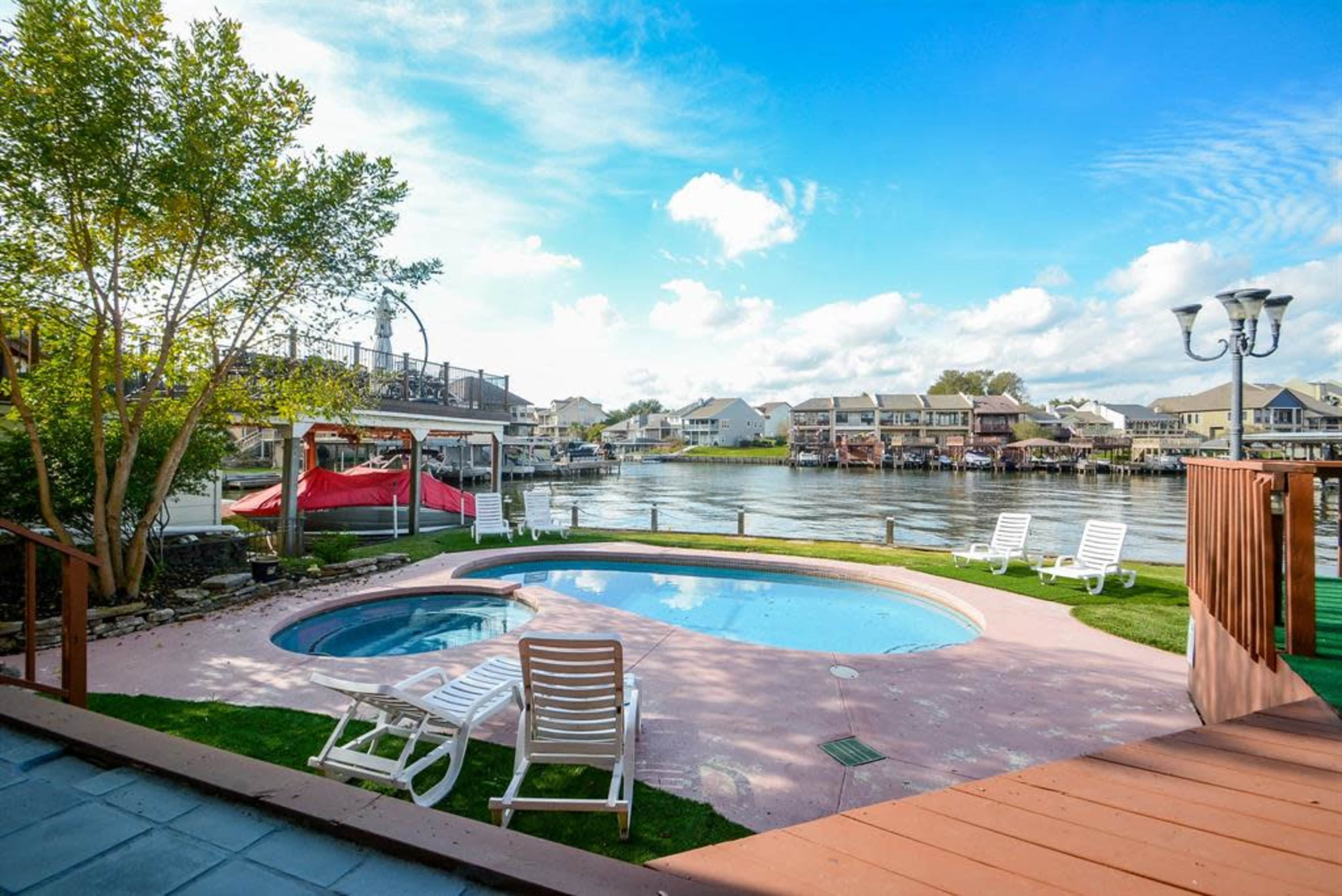 A backyard features a swimming pool and a hot tub overlooking a body of water surrounded by houses.