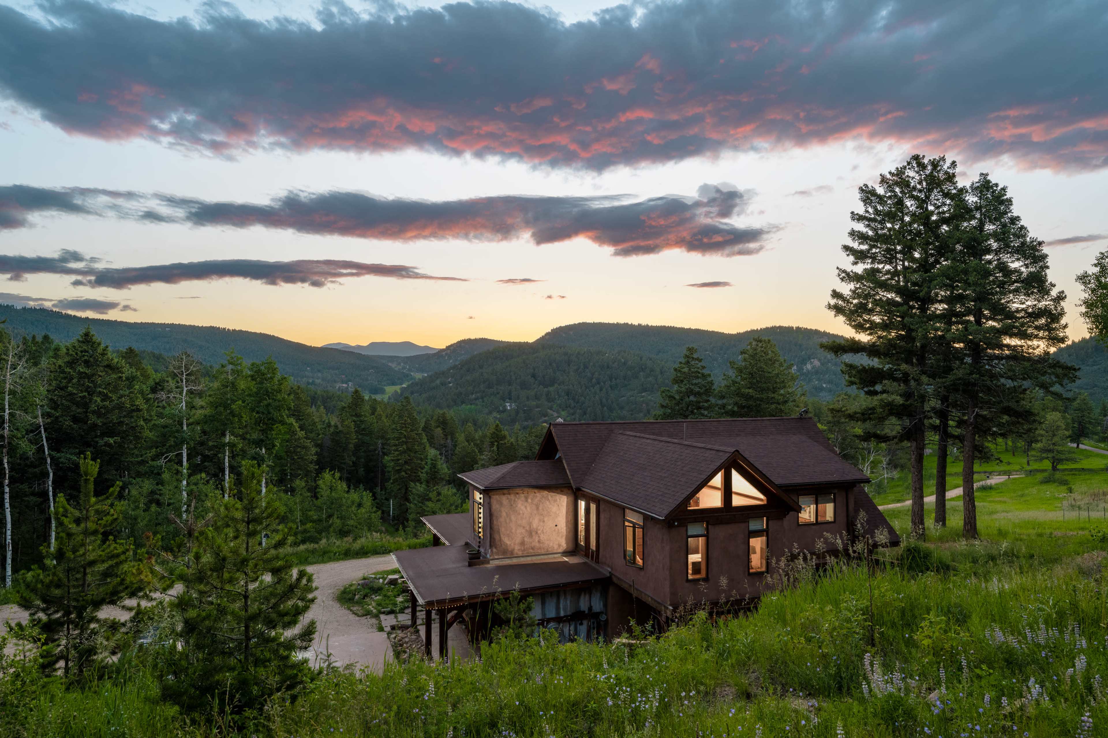 A modern house sits on a hillside, surrounded by trees and rolling mountains under a colorful sunset sky.
