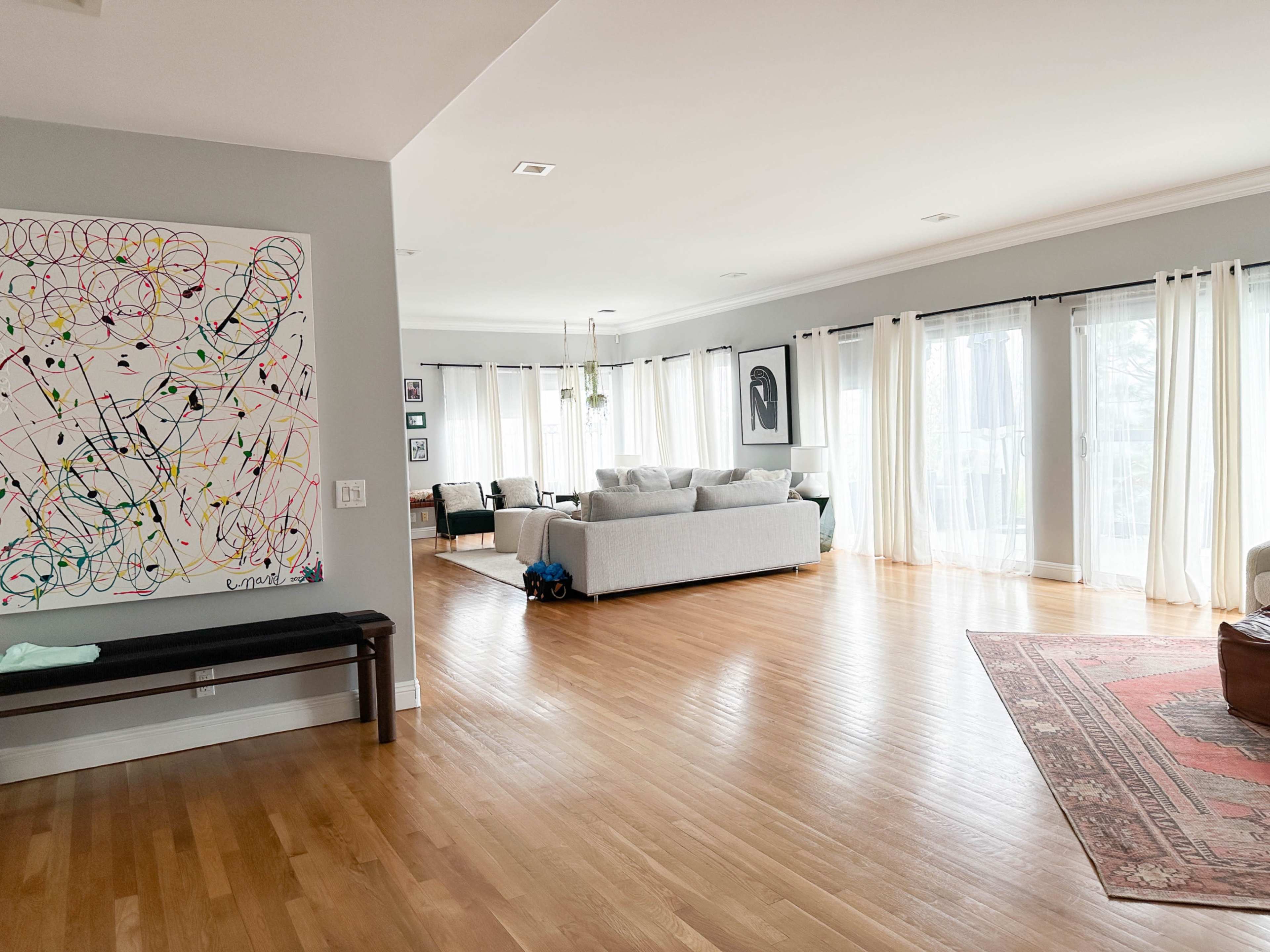 The image shows a spacious living room with large windows, light wood flooring, and contemporary furniture, including a gray sofa and an abstract wall painting.