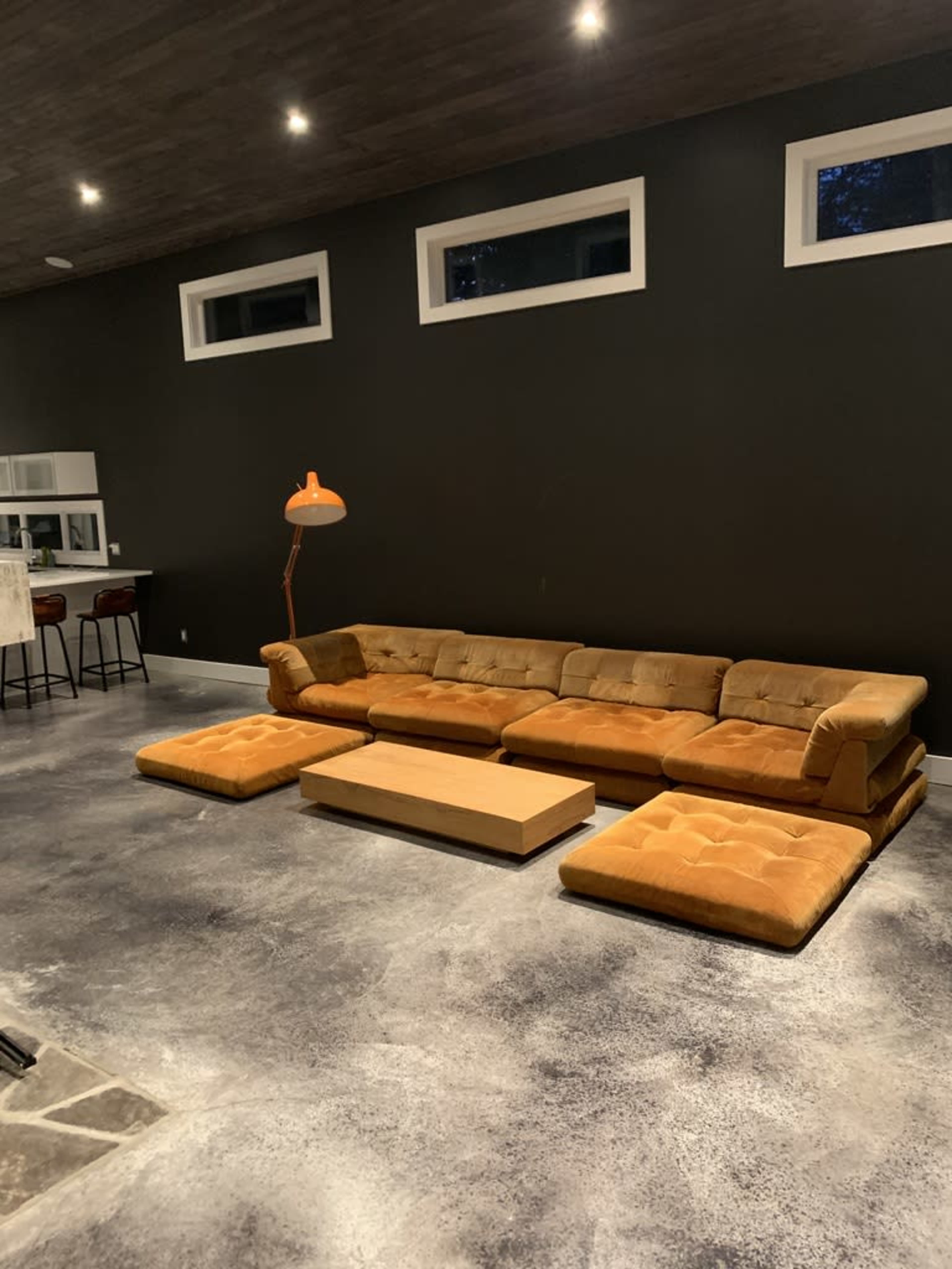 A modern living room with a large, mustard-colored sectional sofa, a wooden coffee table, and a floor lamp, set against a dark wall and concrete flooring.