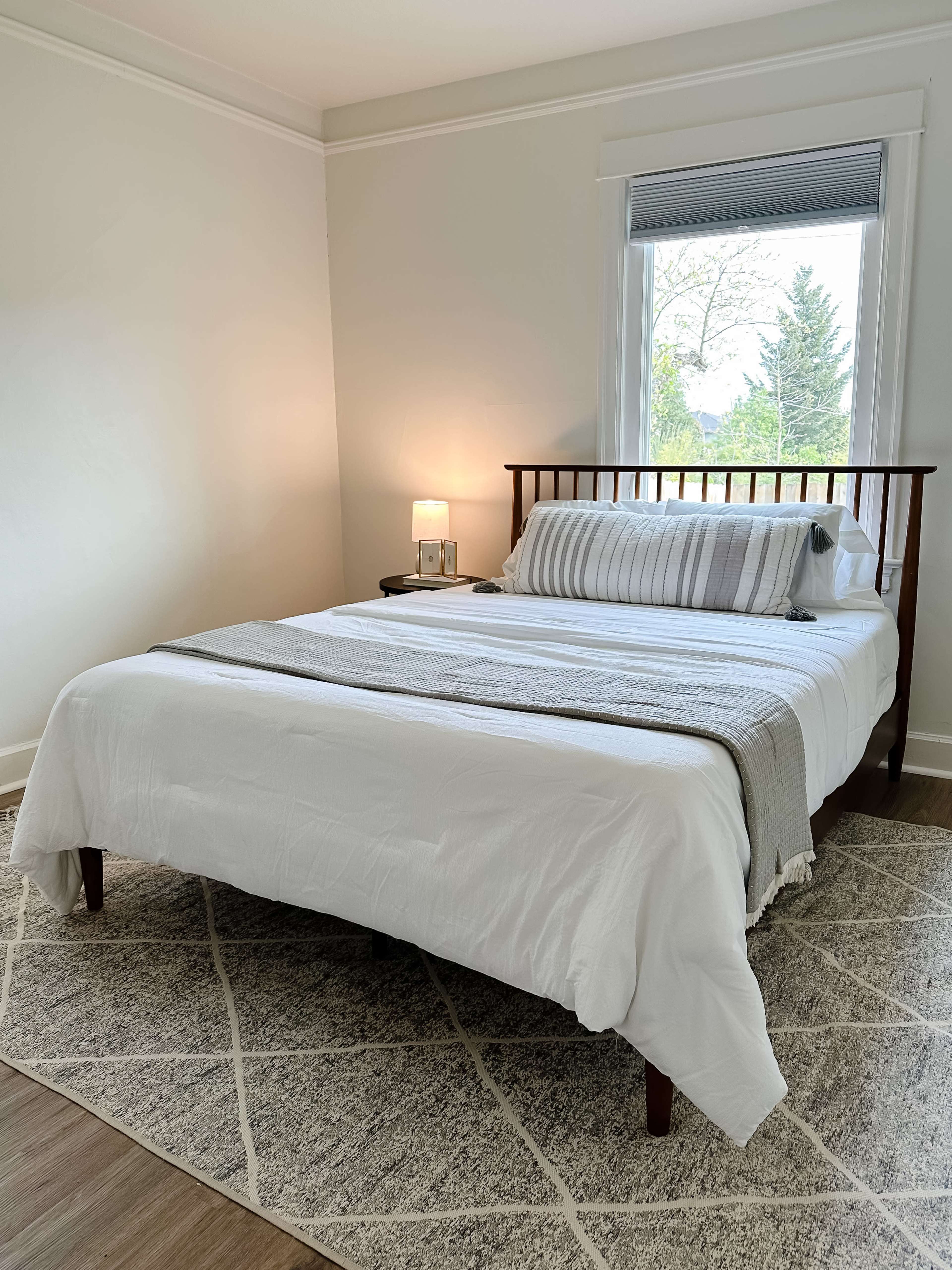 The image shows a neatly made bed in a simple, well-lit bedroom with minimal decor and a window allowing natural light to enter.