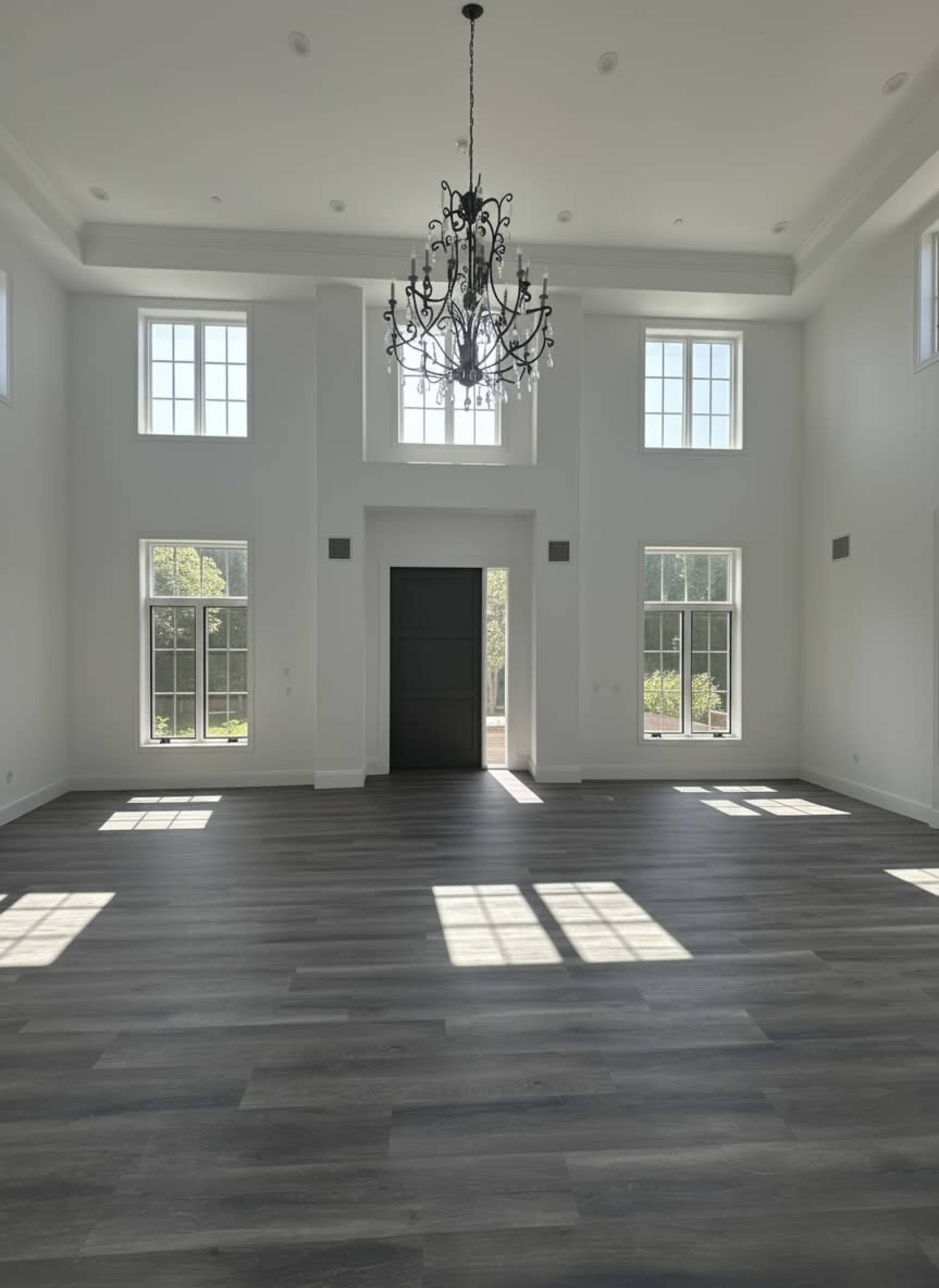 A spacious room with gray wooden flooring, large windows allowing natural light, and a decorative chandelier hanging from the ceiling.