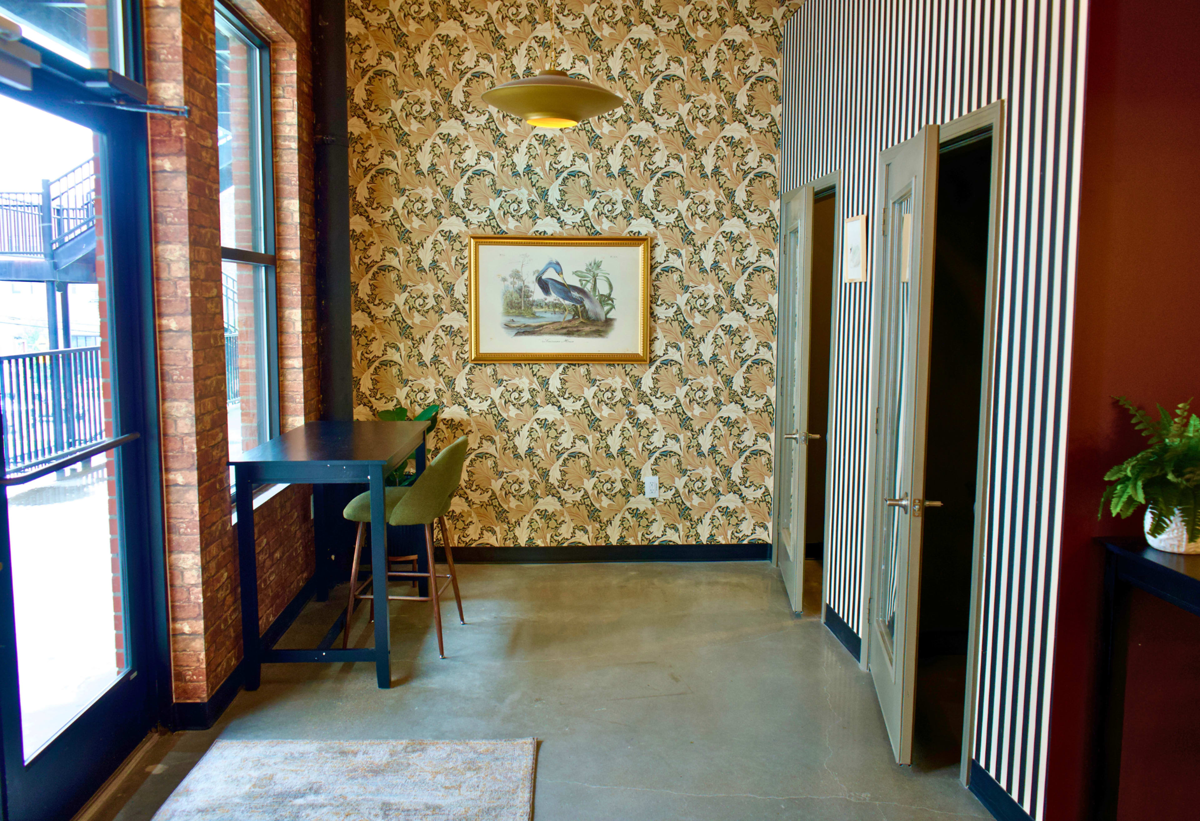 The scene features a small hallway with two doors, a green seating area with a bar-style table, and a decorative wall featuring floral wallpaper and a framed painting.