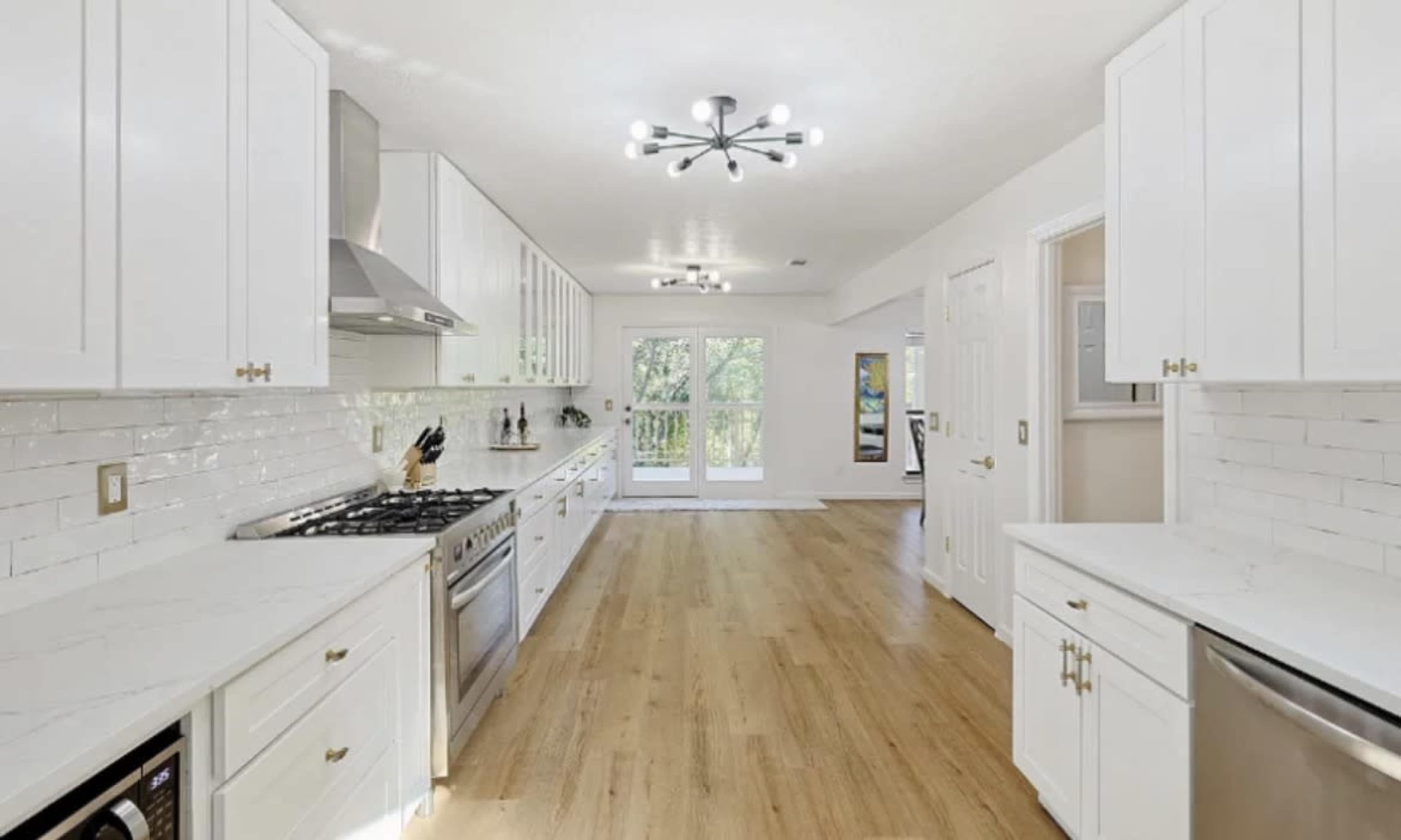The image shows a spacious kitchen with white cabinets, a large gas stove, and hardwood flooring, leading to a set of double doors that open to a view outside.