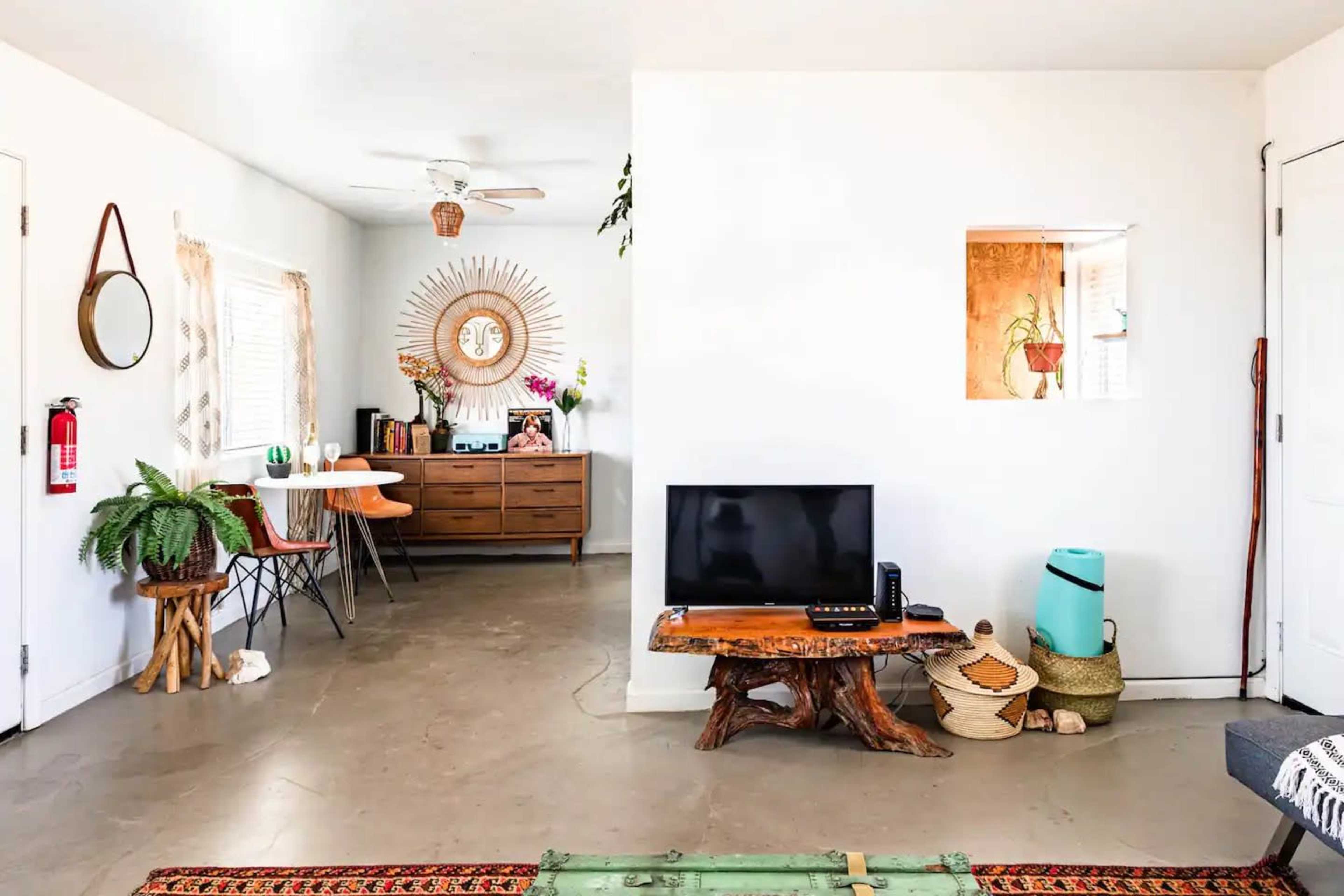 A minimalist living space featuring a wooden table and chairs, a large wall mirror, a TV on a rustic stand, and a small decorative area with plants and textiles.
