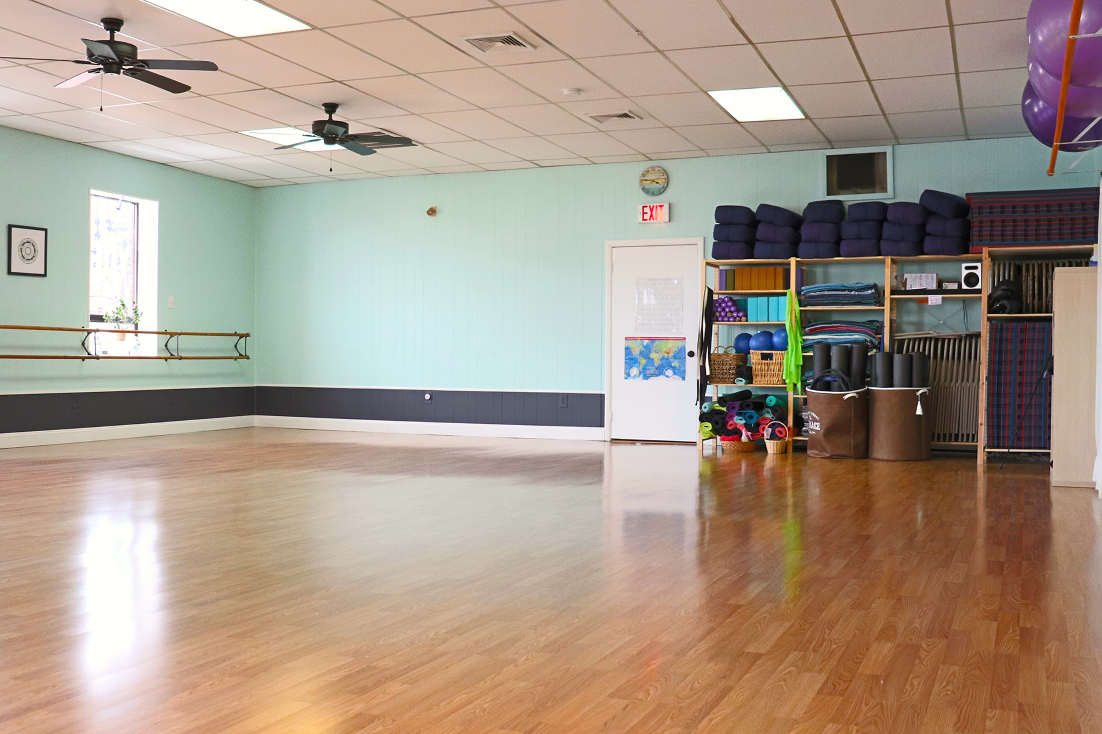 A spacious, empty exercise studio with wooden flooring, mirrors, and storage shelves filled with fitness equipment.