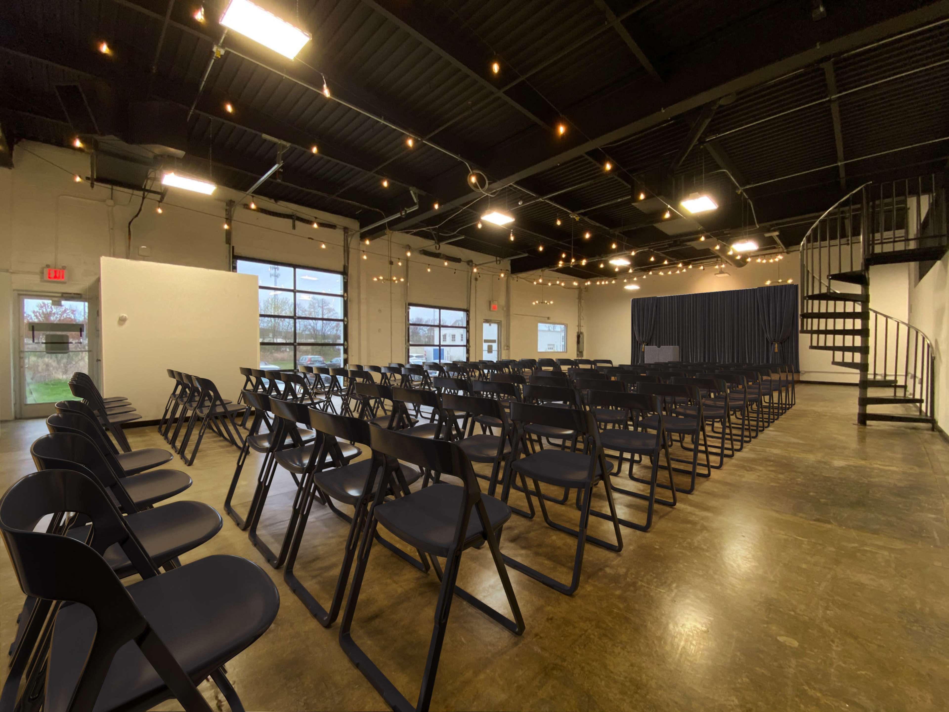 An empty event space with rows of folding chairs arranged facing a stage, complemented by string lights overhead.