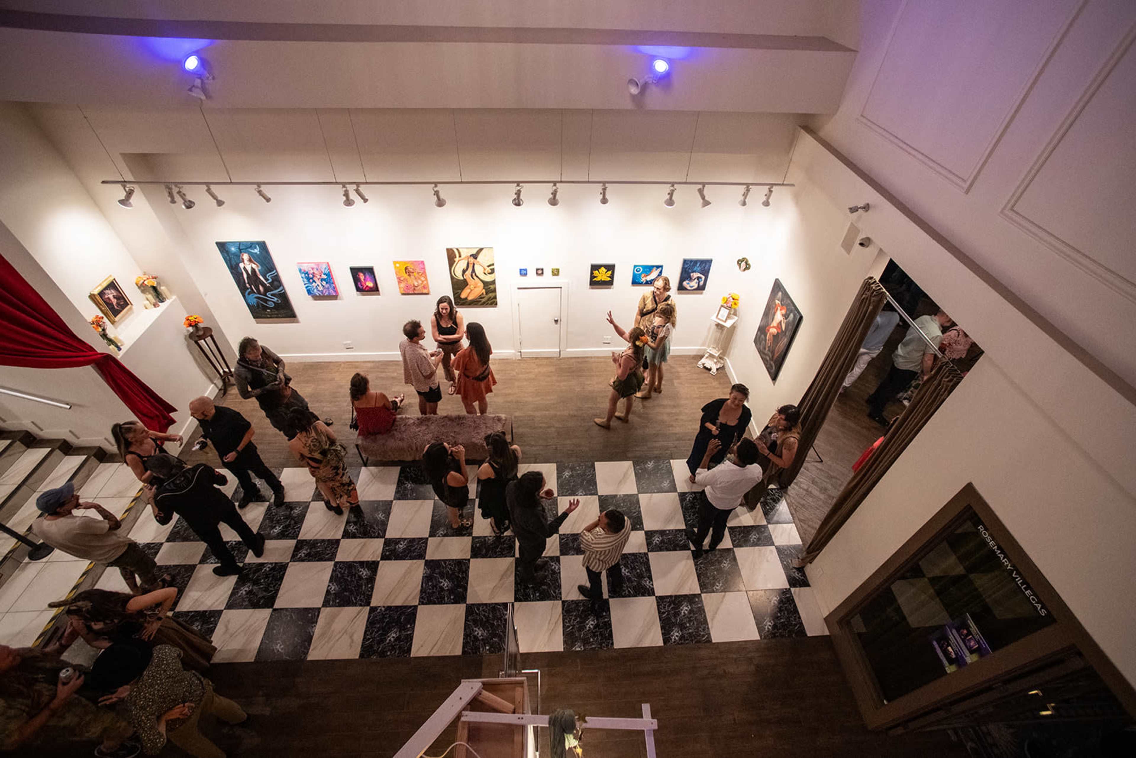 The image shows a lively art gallery opening with attendees mingling and viewing various artworks displayed on the walls.