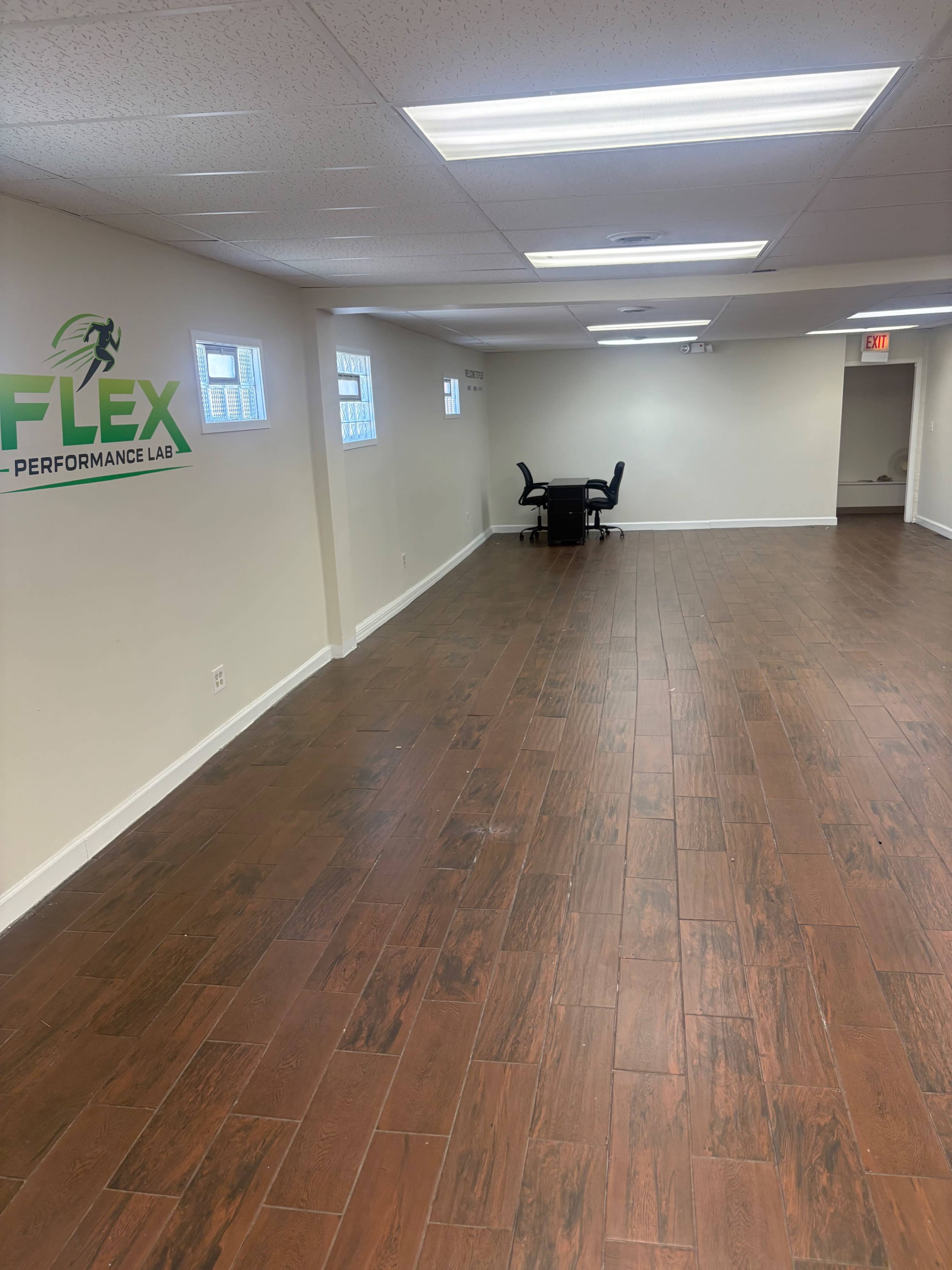 The image shows a spacious, empty room with wooden flooring and a desk with two chairs in one corner.